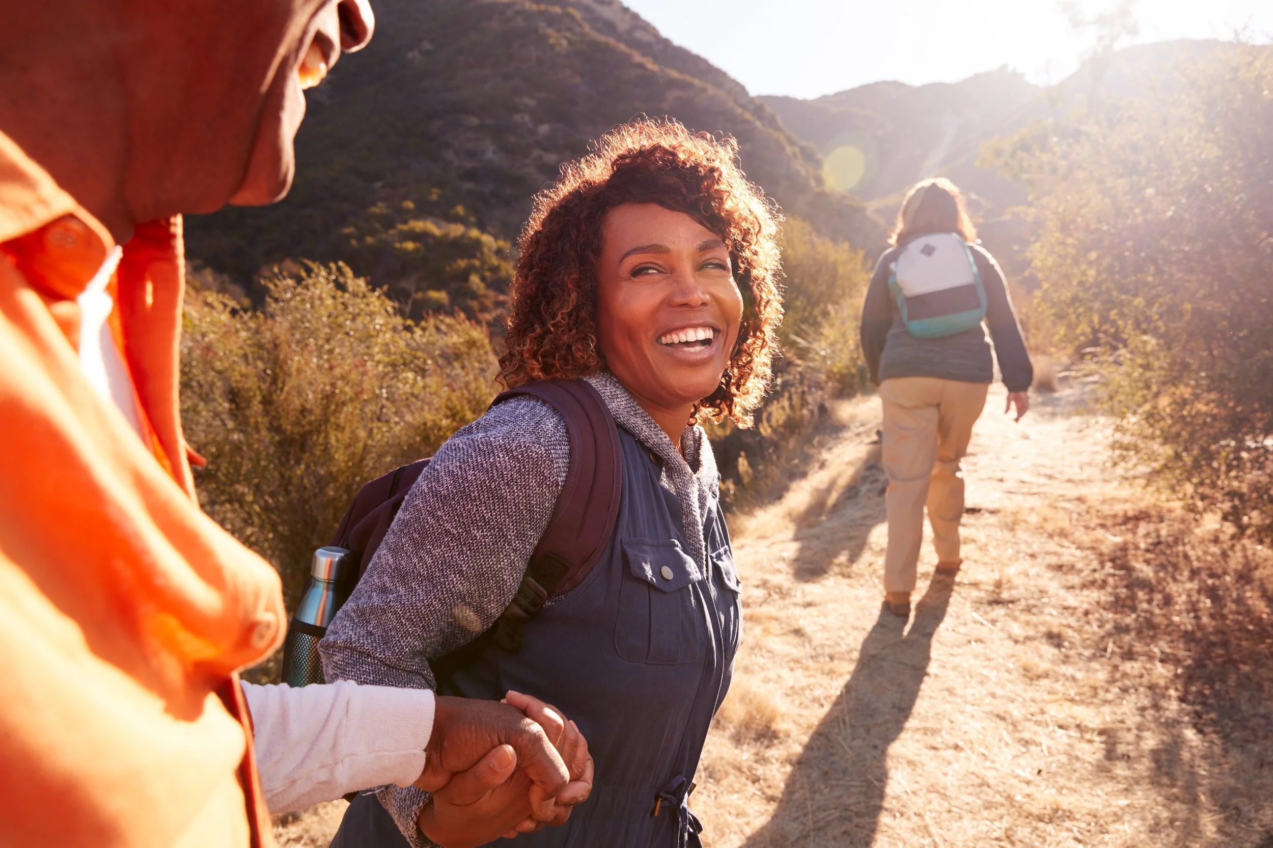 Happy woman with curly hair smiling and holding hands with a person in an orange jacket during a hike on a trail in a mountainous area
