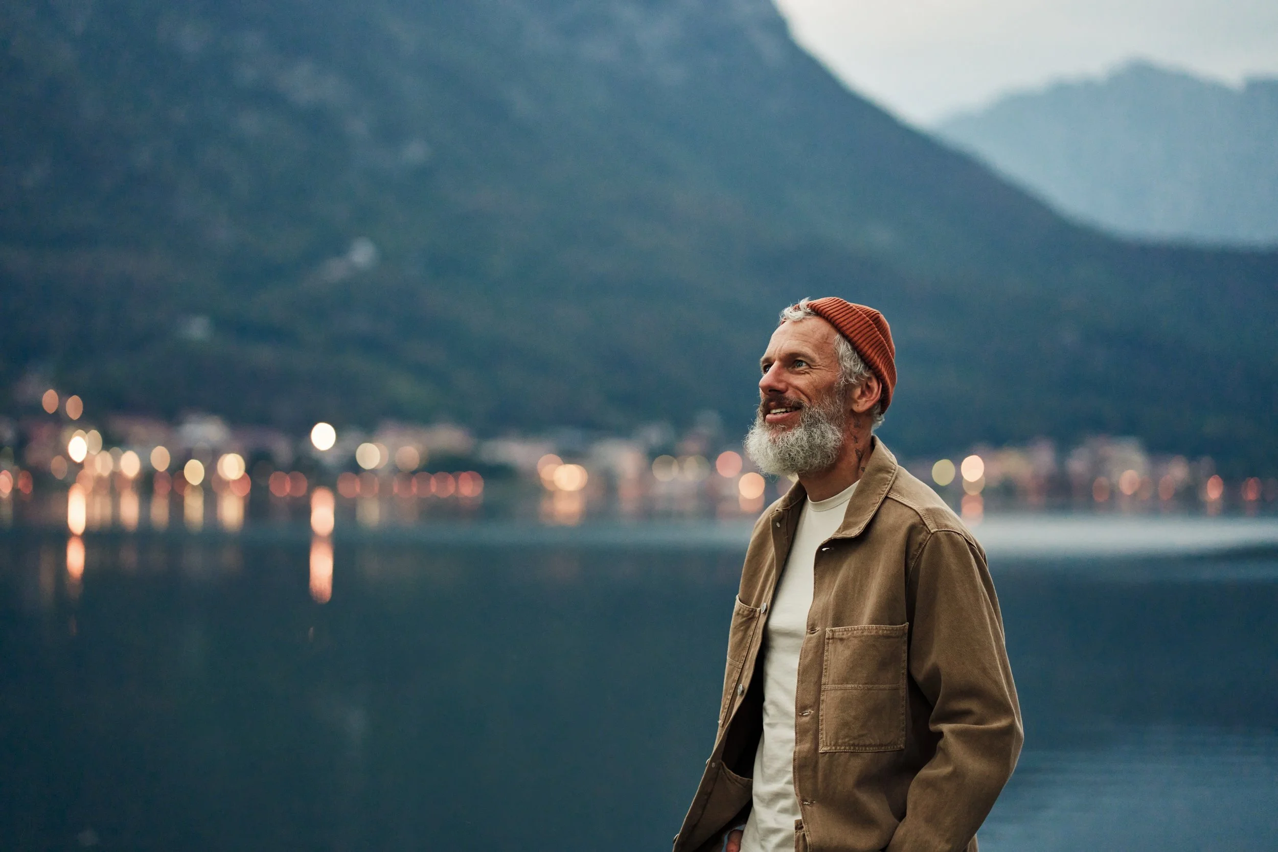 A man with a gray beard and mustache, wearing a brown jacket, a white shirt, and a red beanie, standing outdoors near a body of water with mountains and city lights in the background at dusk.