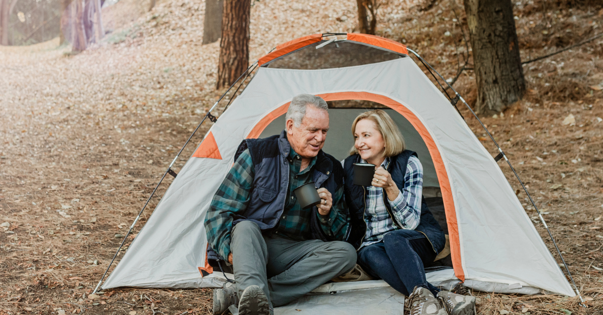 An elderly couple sits inside a tent in a forest, smiling and enjoying coffee or tea.
