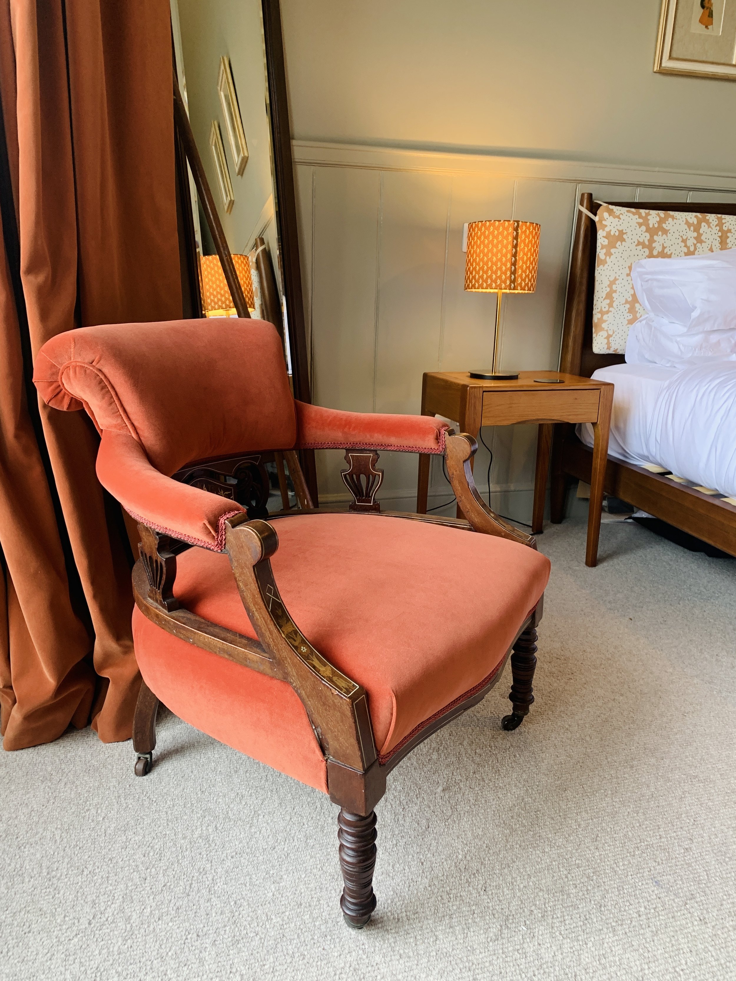 Vintage pink velvet armchair with carved wooden legs and armrests in a cozy bedroom, next to a wooden side table with a yellow patterned lamp, positioned near a bed with white linens and a decorative headboard.