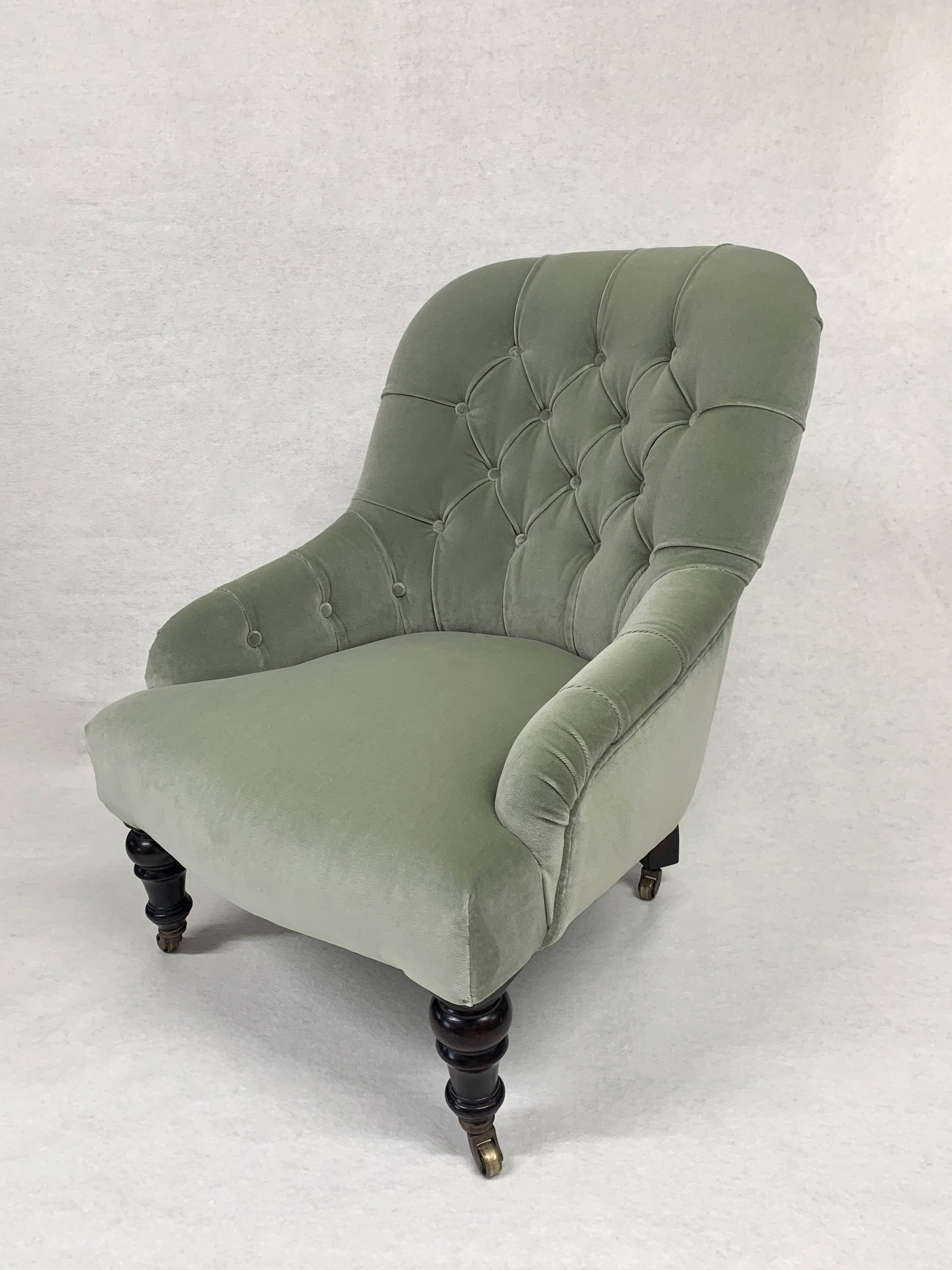 A vintage green upholstered armchair with tufted backrest and dark wooden legs with small brass casters, set against a plain light-colored background.