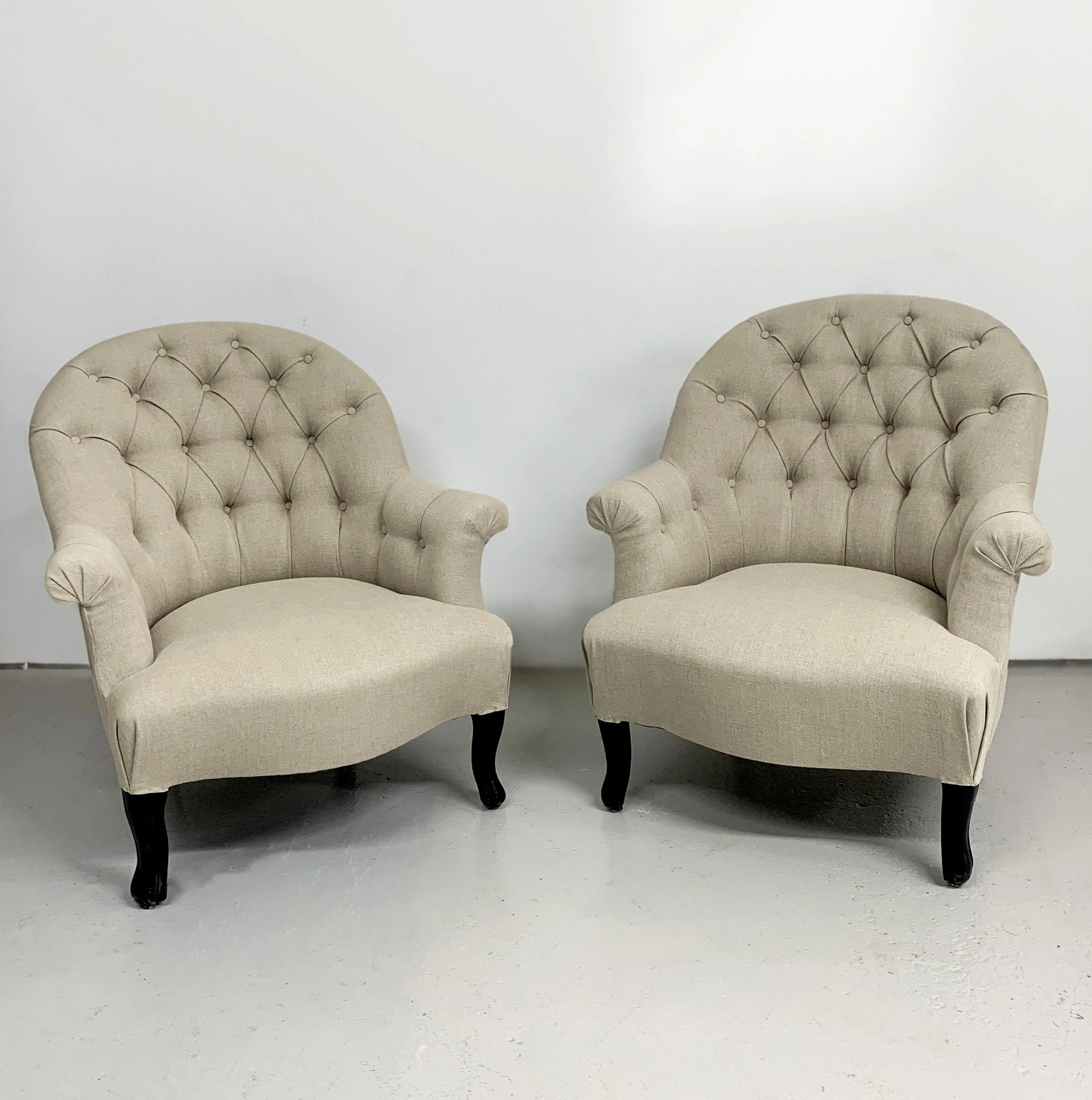 Two beige upholstered armchairs with tufted backs, rolled arms, and dark curved wooden legs, placed side by side against a plain white wall.