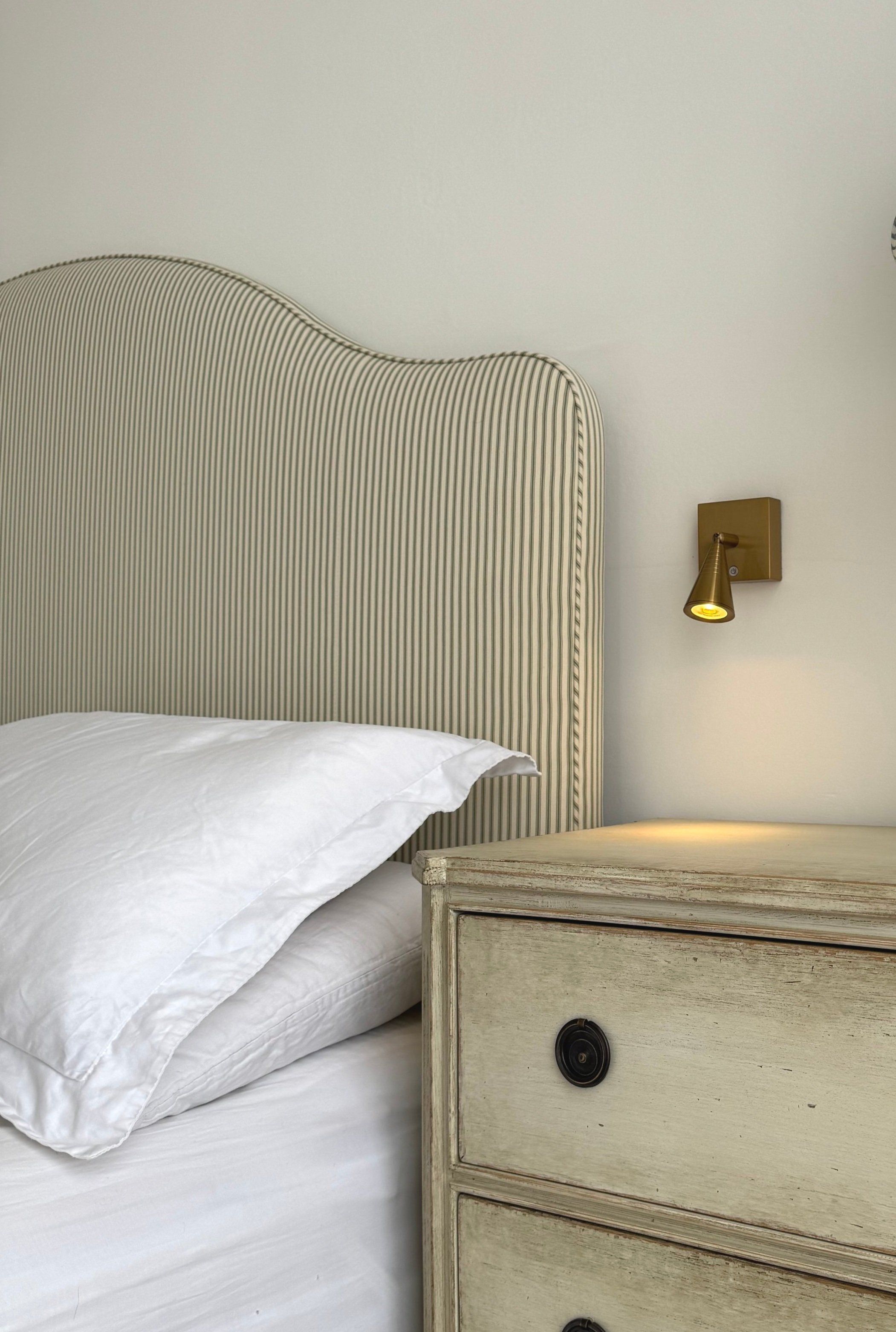 A close-up of a bedroom corner showing a beige striped headboard, a white pillow, a white bed sheet, a wooden nightstand, and a wall-mounted reading light.