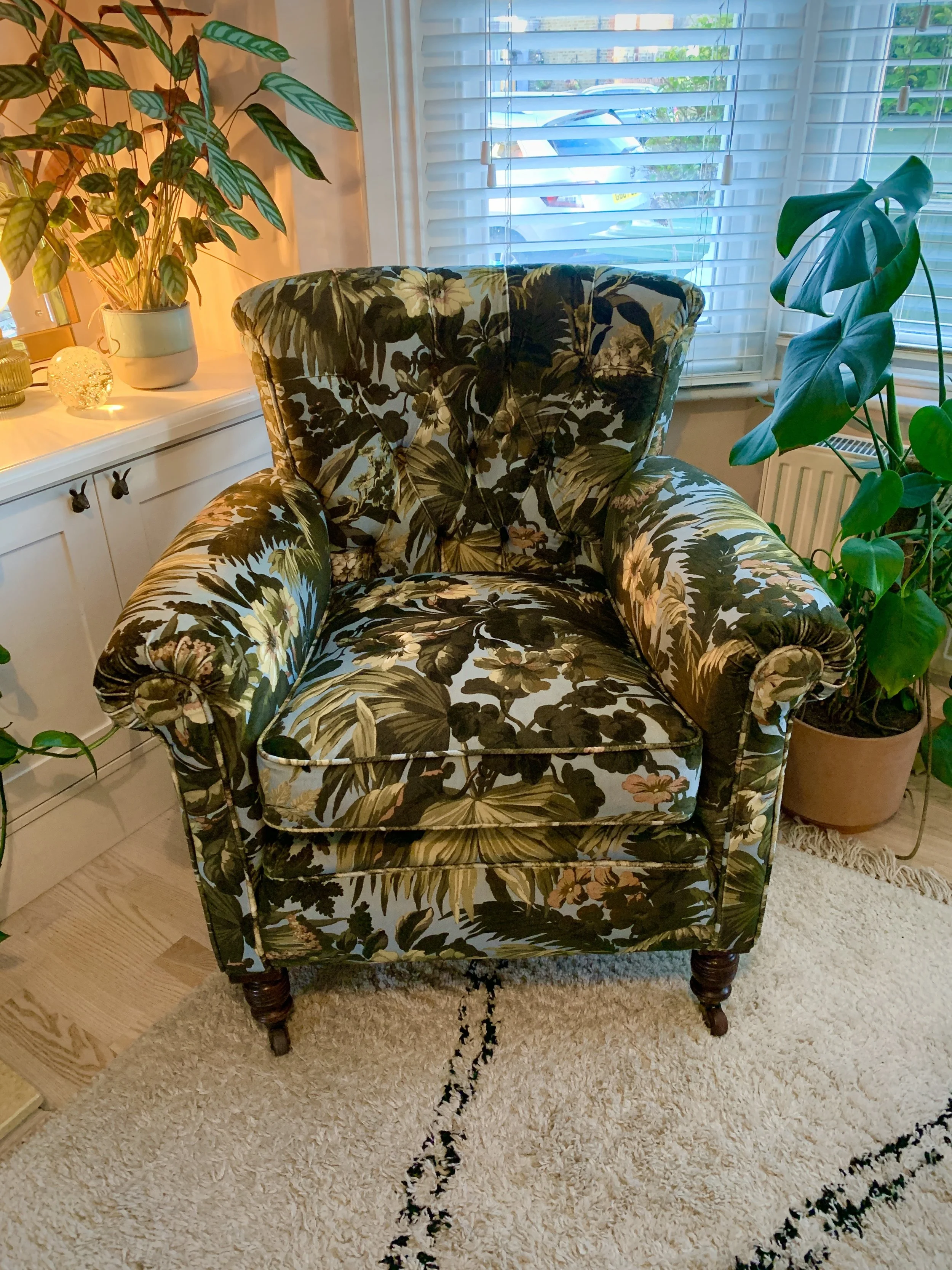 A vintage armchair with a floral and foliage patterned upholstery, placed in a cozy living room corner near a window and surrounded by potted houseplants.