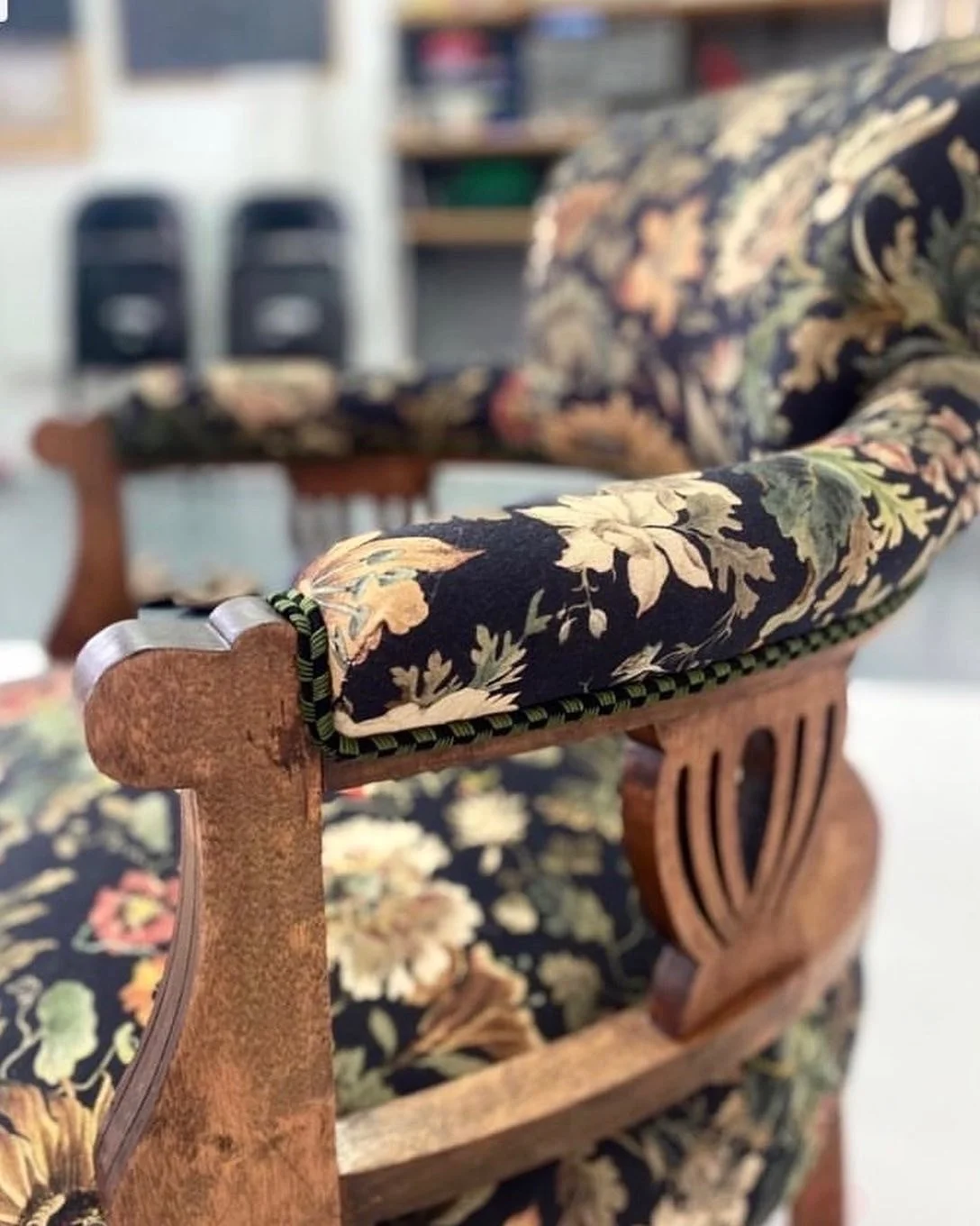 Close-up of a wooden chair with floral upholstered cushion and backrest, in a room with shelves and speakers in the background.
