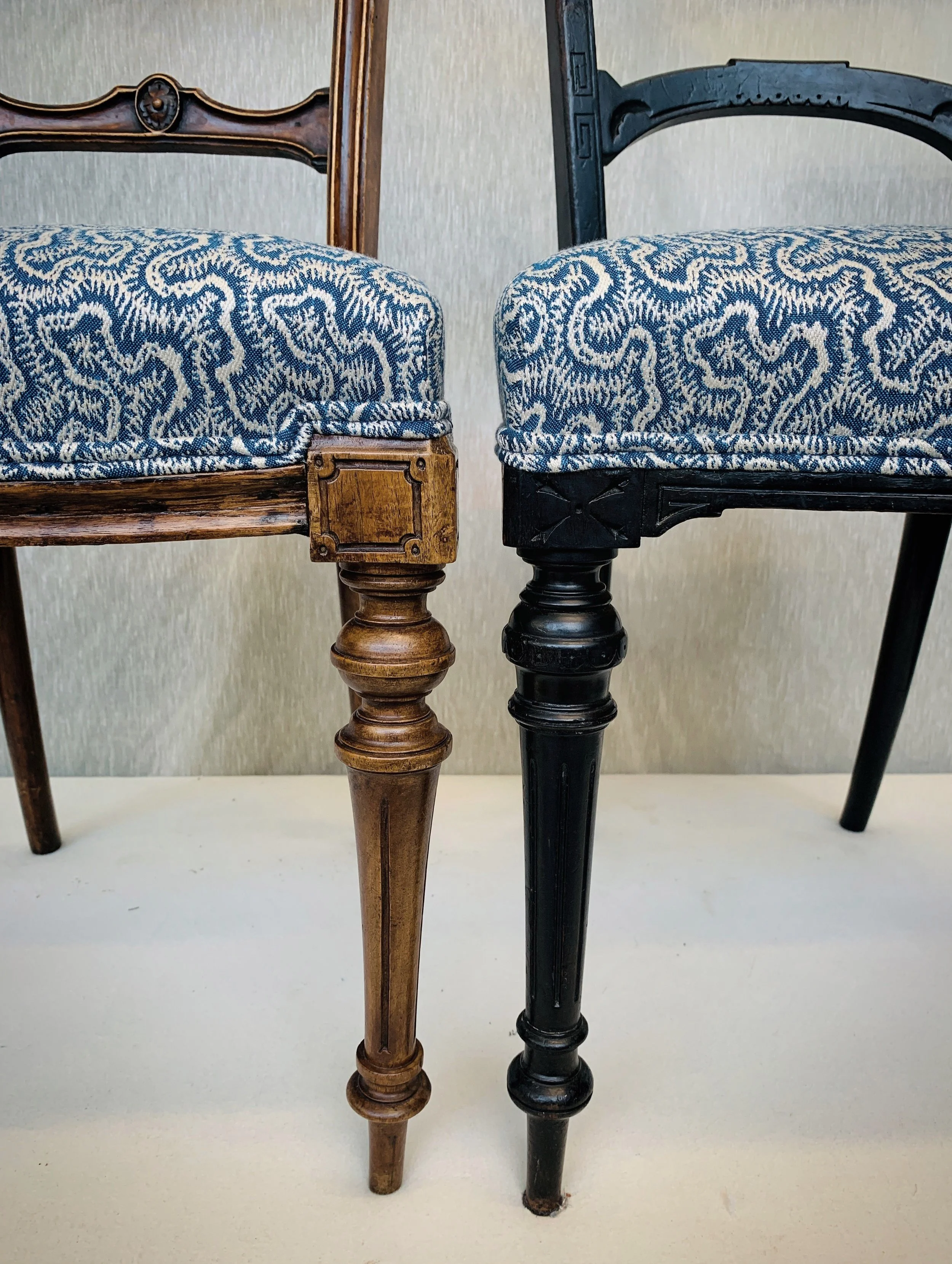 Close-up of two vintage chairs with patterned fabric seats, one with a gold-colored wood frame and the other painted black, showing their ornate carved legs and backs.