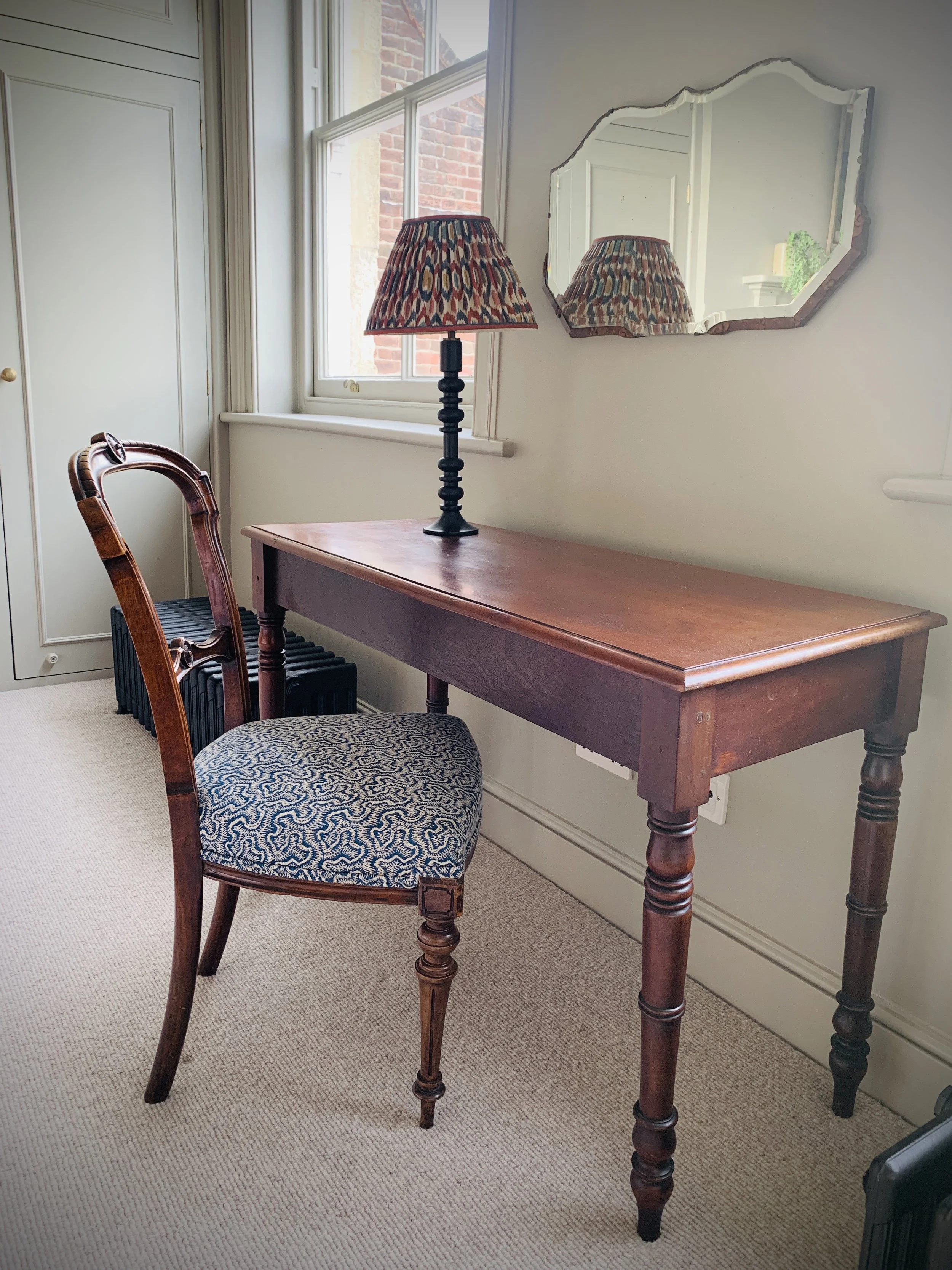 A wooden desk with a patterned glazed lampshade on a lamp, a chair with a patterned upholstered seat, a window with brick walls outside, and a wall mirror reflected in the frame.