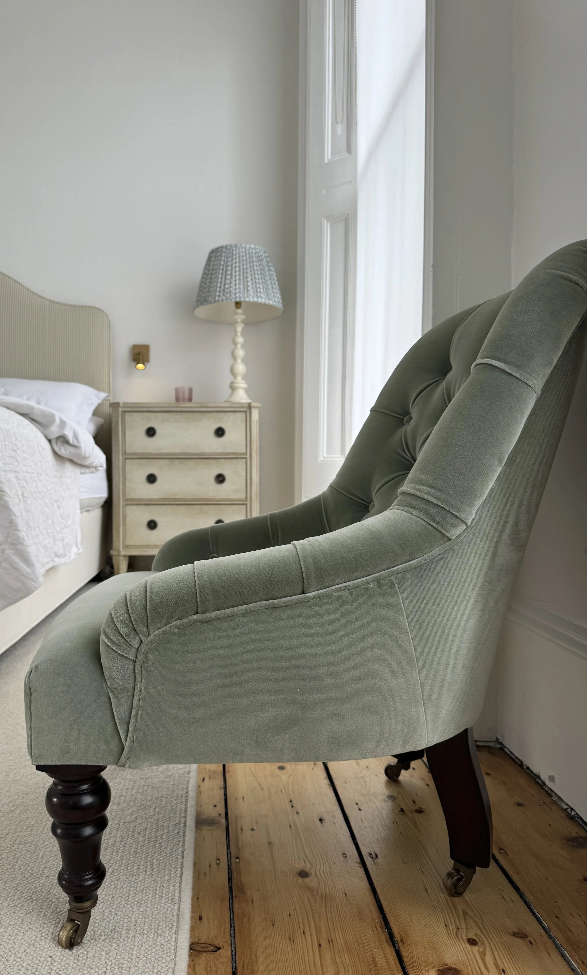 A light green upholstered armchair with wooden legs on wheels is placed next to a bed with a beige headboard and white bedding. A small wooden nightstand with three drawers, a blue-patterned table lamp, and a small pink candle are in the background. There is a white wall and a window with white curtains to the right.