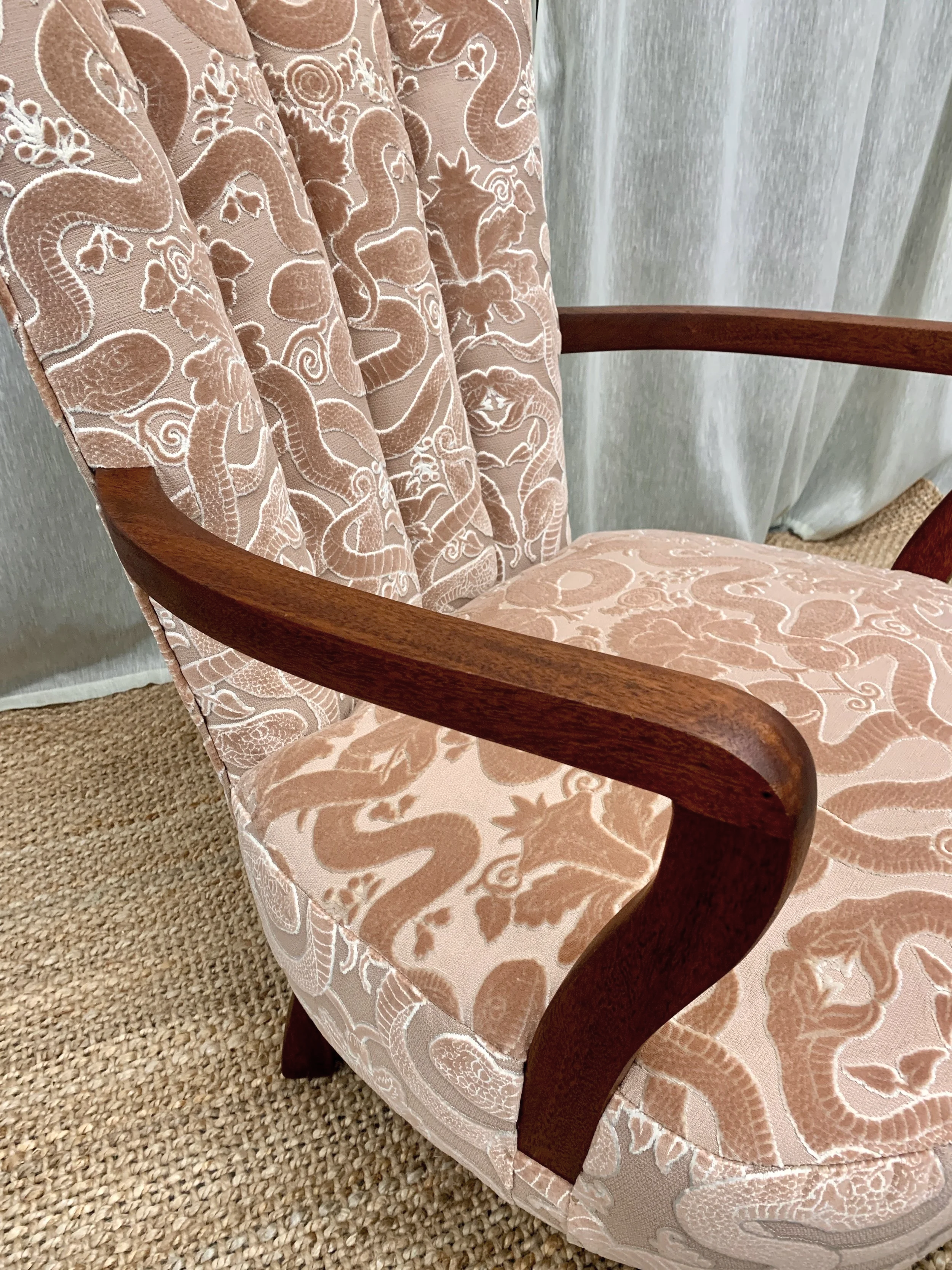 Close-up of an elegant upholstered armchair with a pink fabric featuring embroidered snake and floral patterns, wooden armrests, and a beige textured carpet beneath.