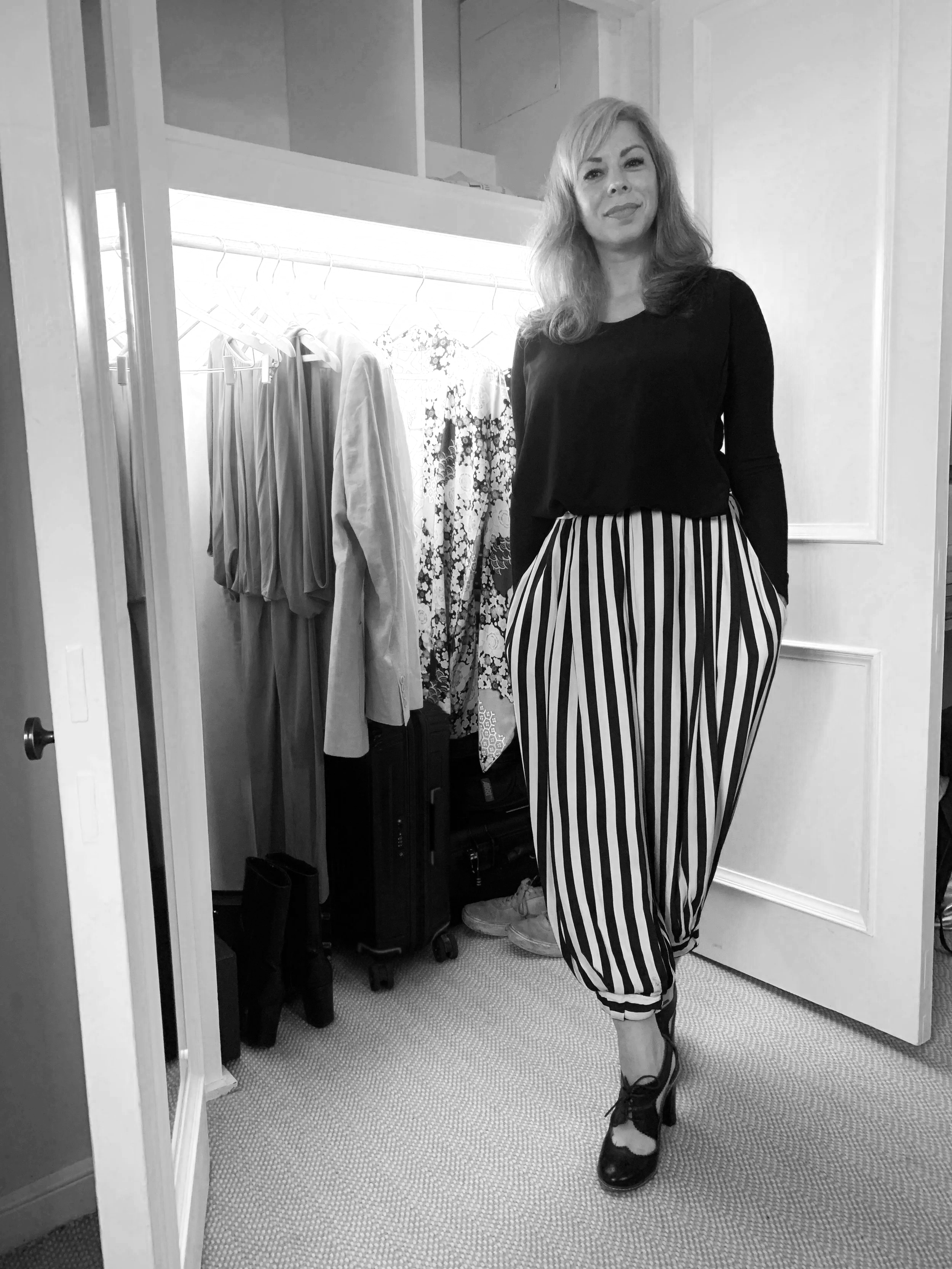 A woman with wavy blonde hair standing in a closet wearing a black long-sleeve top and striped black and white loose-fitting pants, with her hands in her pockets.