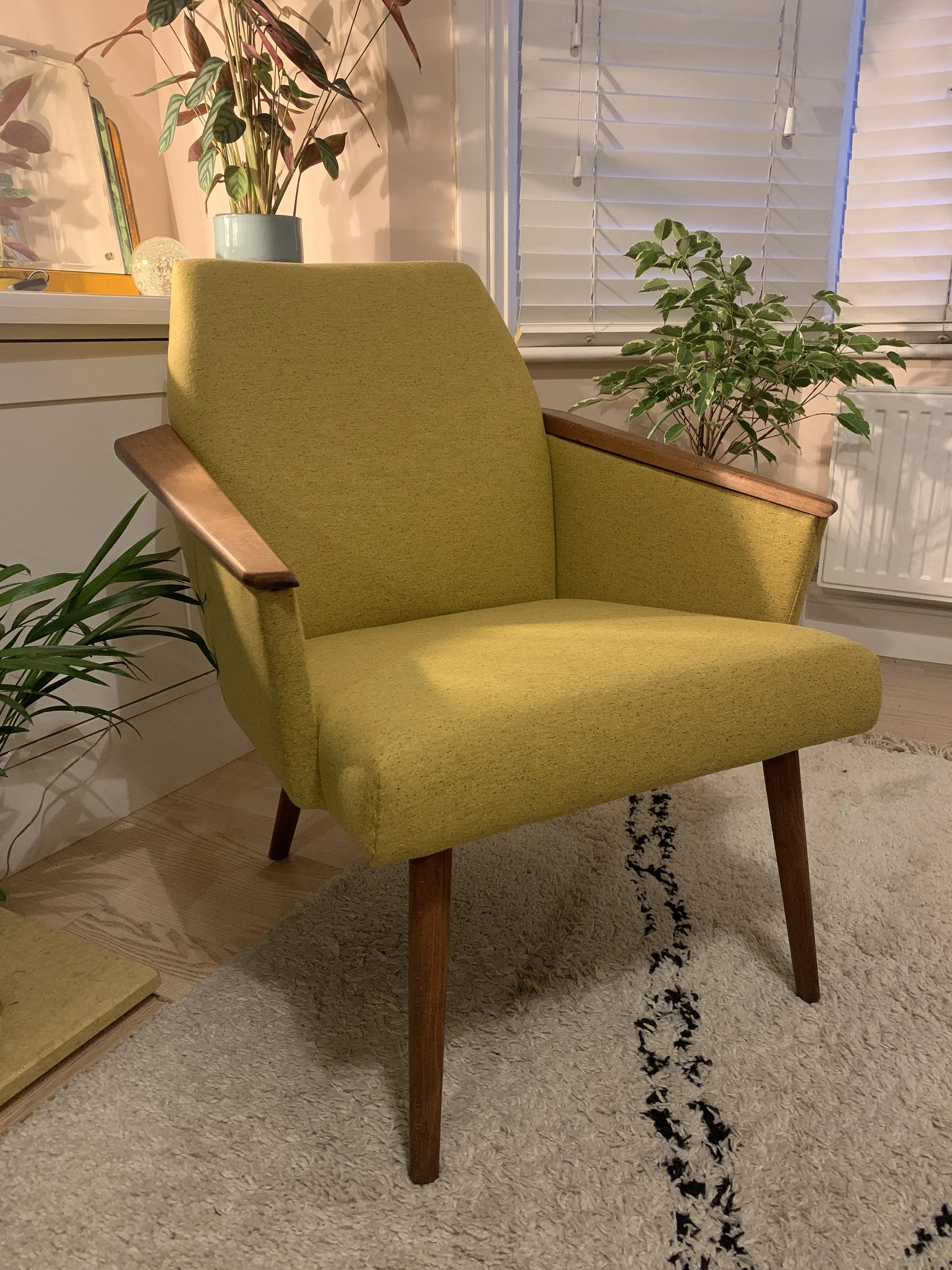 A mid-century modern green armchair with wooden armrests and legs, situated in a cozy living room corner with houseplants, a window with blinds, and a beige rug.