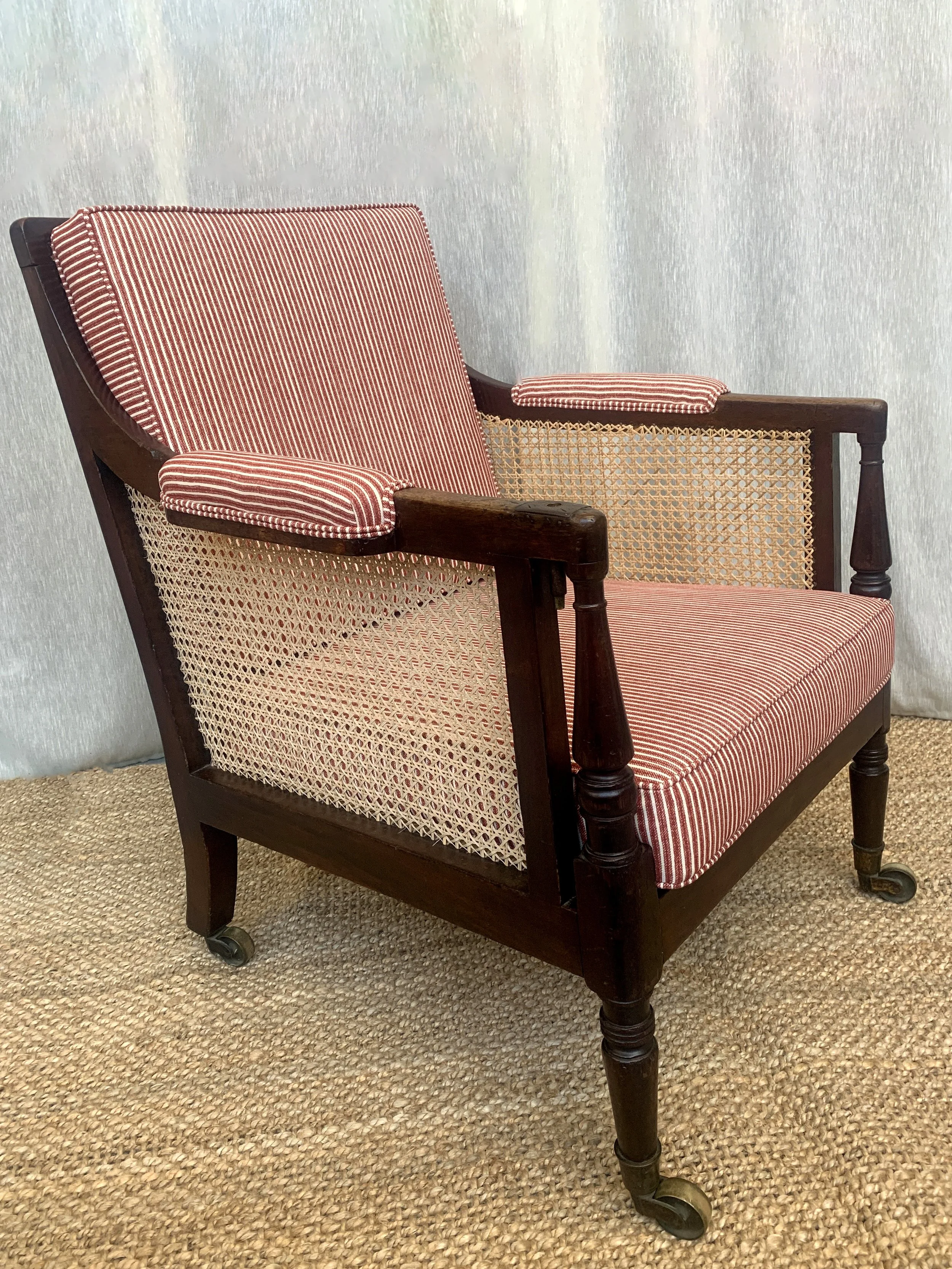 Vintage armchair with red and white striped cushions, wooden frame, cane sides, and small casters on a woven rug.