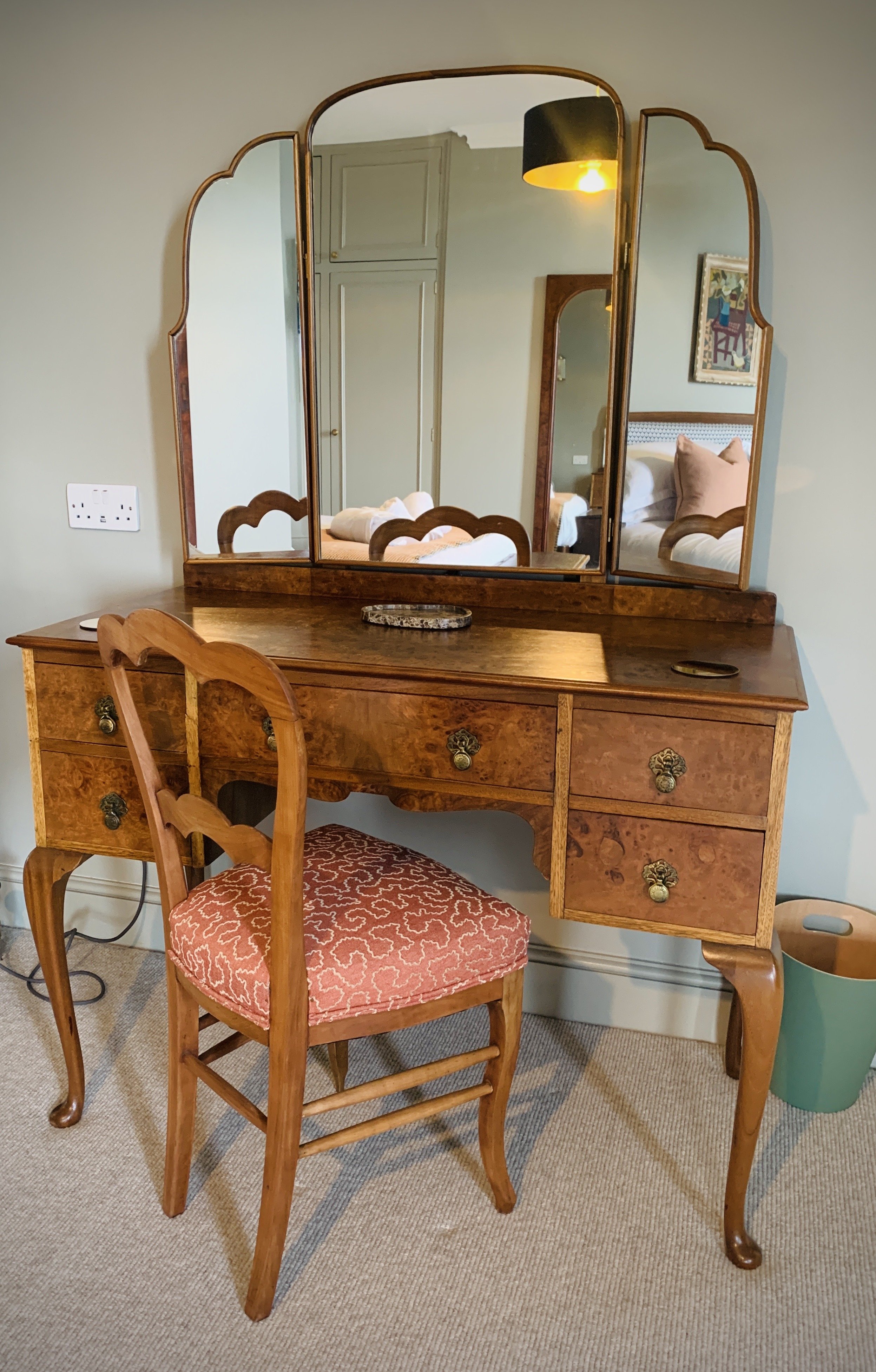 A vintage wooden dressing table with a large mirror and a matching chair, positioned in a bedroom with beige carpet and light green walls. The mirror reflects part of a bed with pillows and a framed picture.