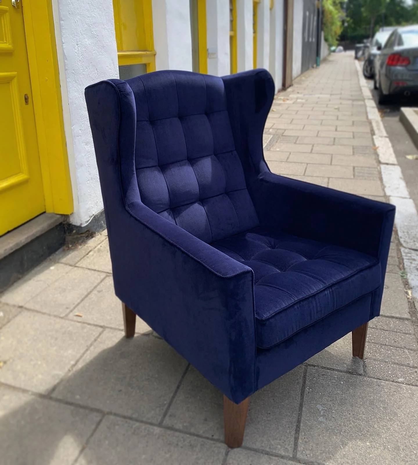 A royal blue vintage armchair with tufted upholstery and wooden legs, placed on a city sidewalk near a yellow door and parked cars.