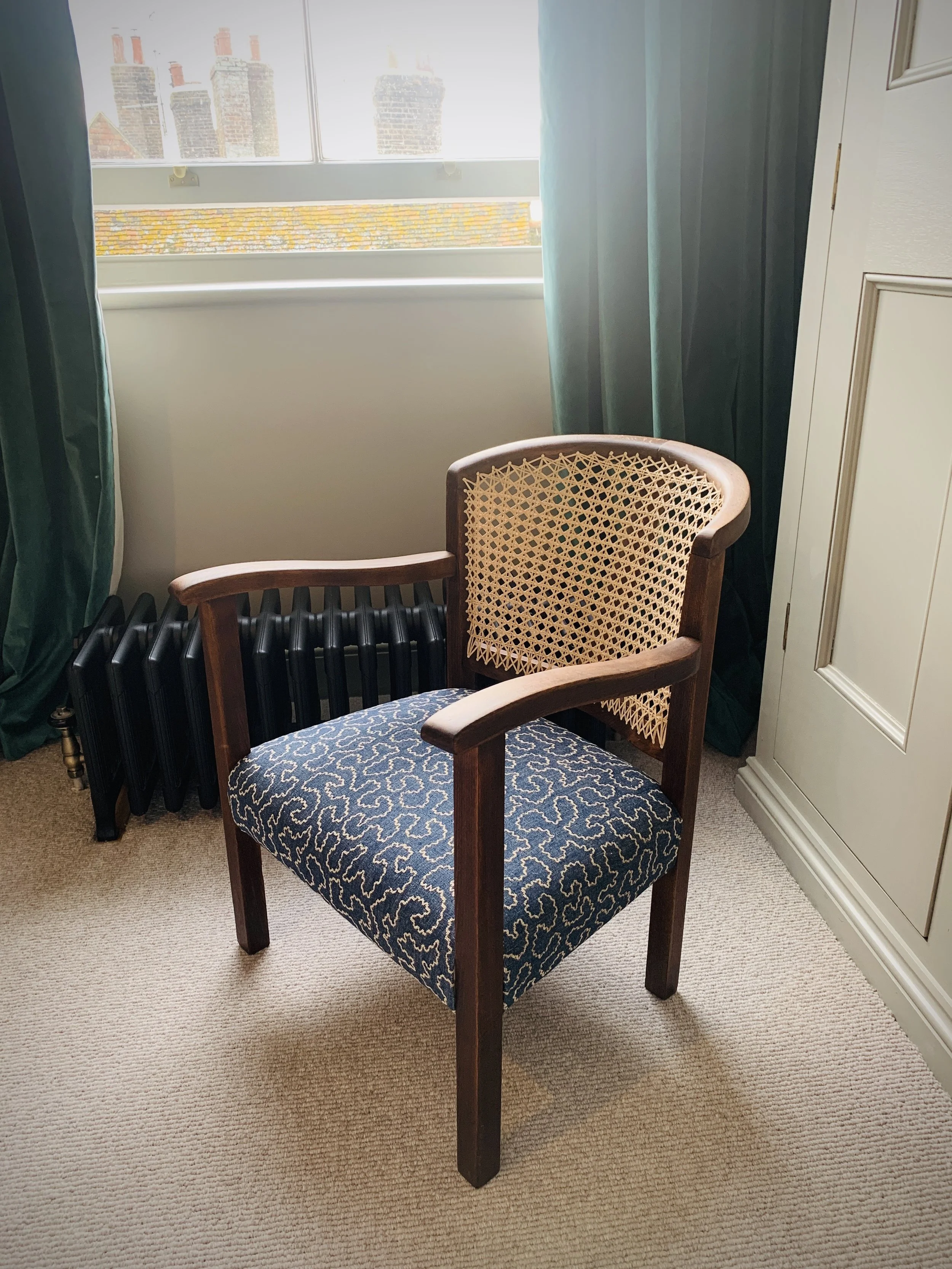 A wooden armchair with a woven cane back and a Scandinavian-style upholstered seat in dark blue with beige patterns. The chair is placed on a beige carpet near a window with green curtains, a black radiator, and a cream-colored cabinet.