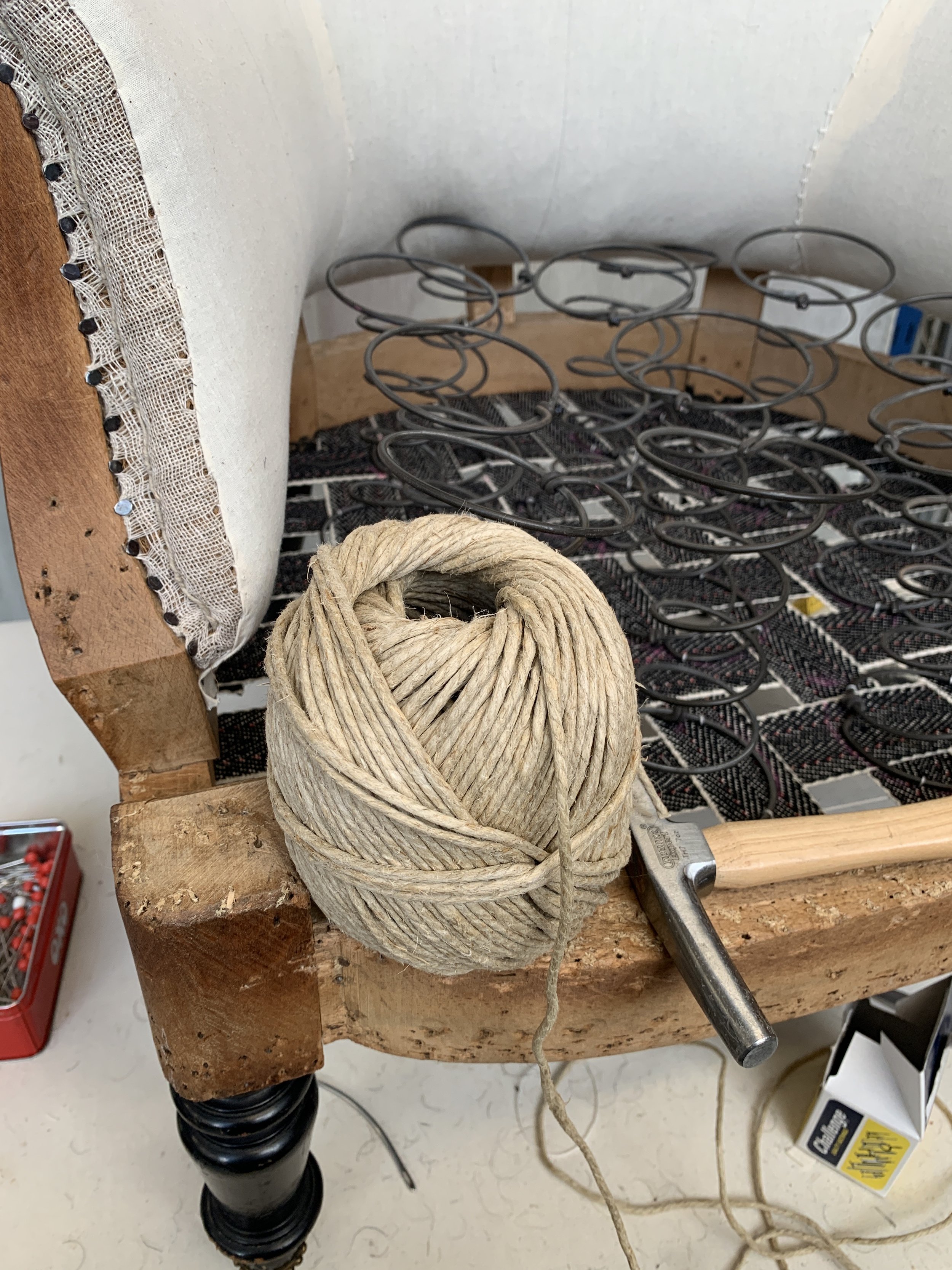 A ball of beige twine rests on a wooden workbench next to a hammer, with a partially completed chair frame and coiled metal springs in the background.