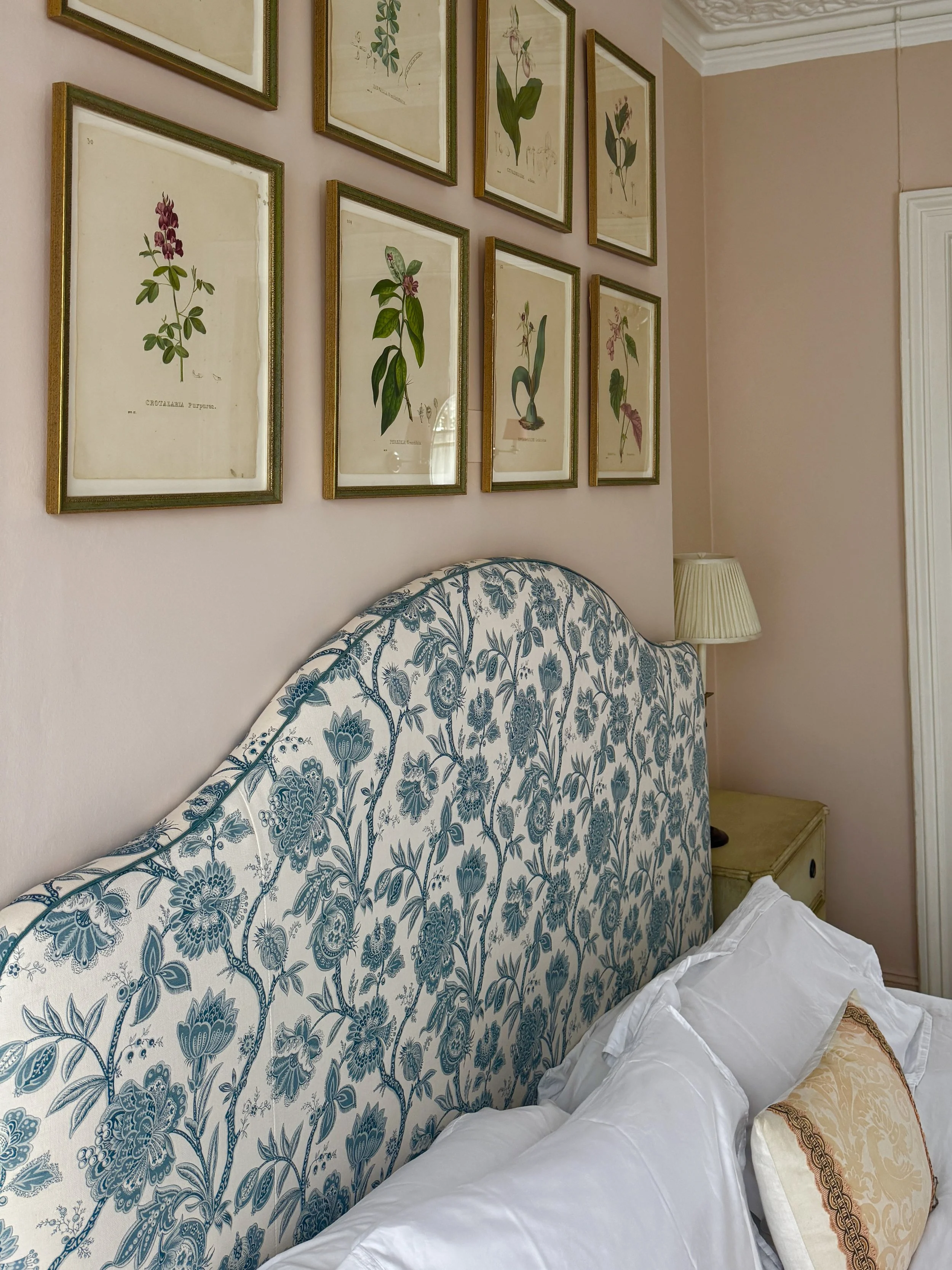 A bedroom with a blue floral-patterned upholstered headboard, white bedding, and a beige decorative pillow. Above the bed, there are several framed botanical illustrations arranged on a pink wall, and to the side, a cream-colored table lamp on a nightstand.