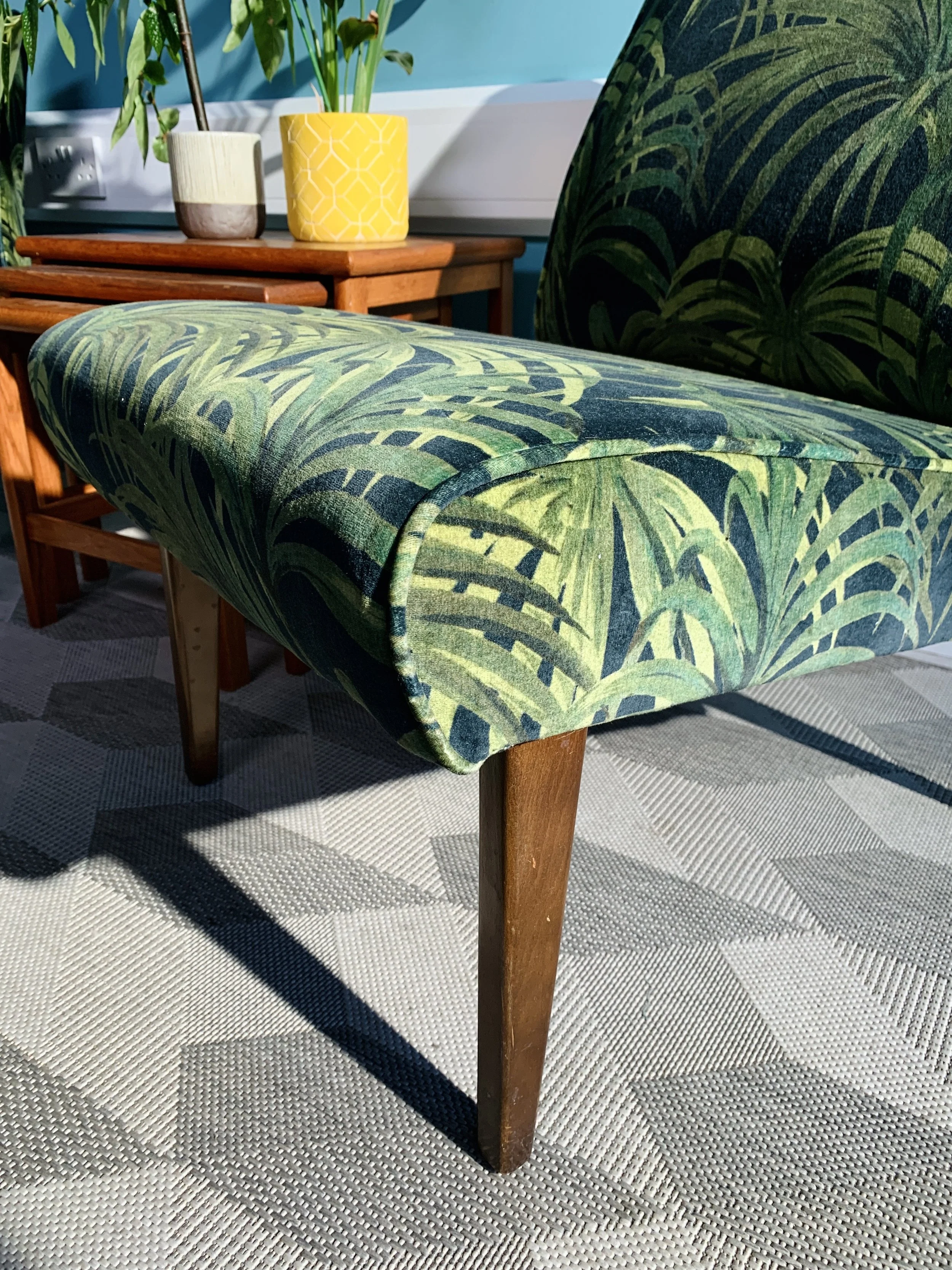 A close-up of a vintage-style green upholstered bench with a tropical leaf pattern, wooden legs, placed on a light-colored textured rug in a room with plants and decorative items on a wooden side table.