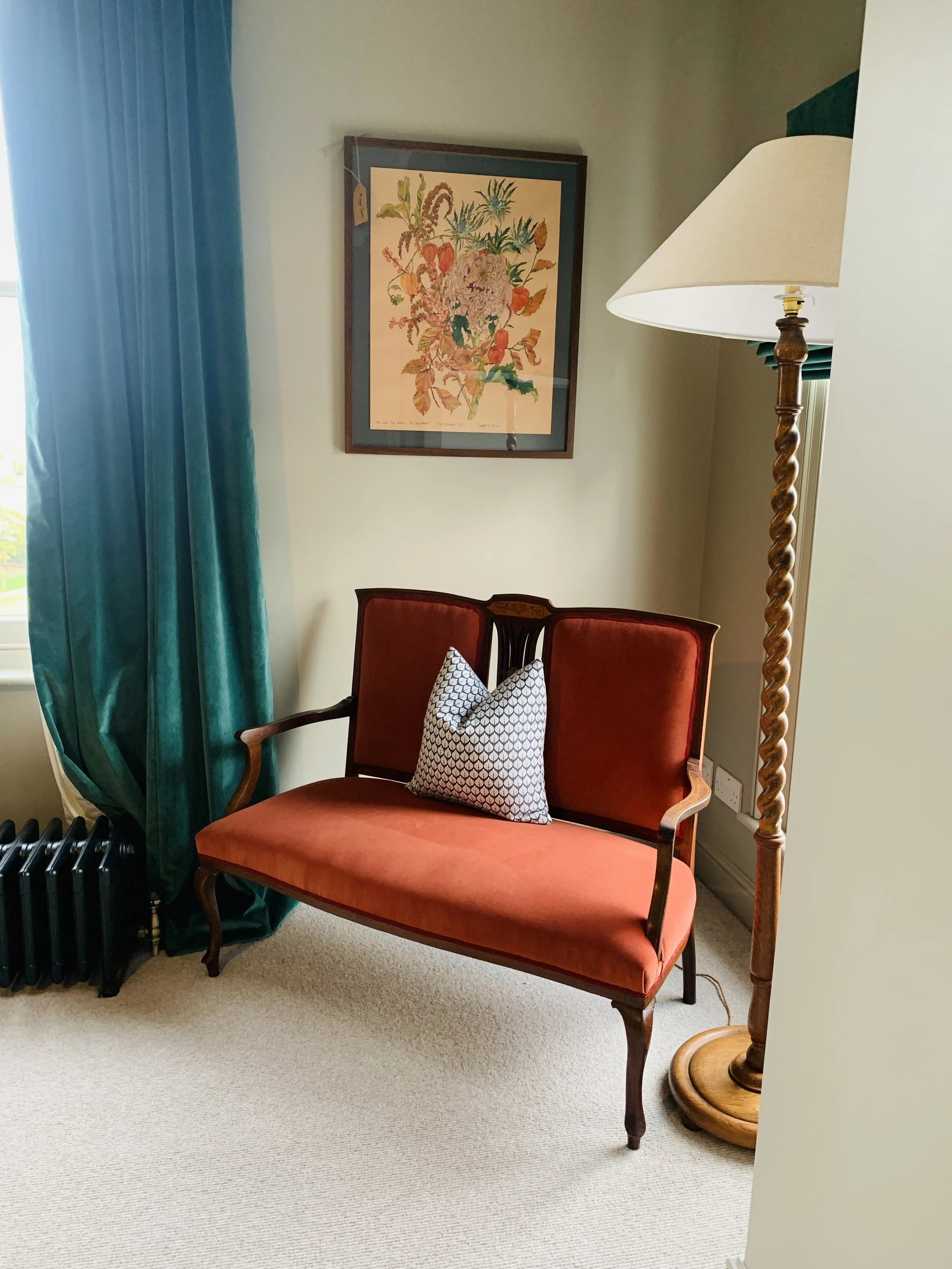 A vintage-style red velvet and wood settee with curved armrests, a small geometric patterned pillow, a framed floral painting above, teal curtains to the side, a standing lamp with a spiral wood post, beige carpet, and a window with a view outside.