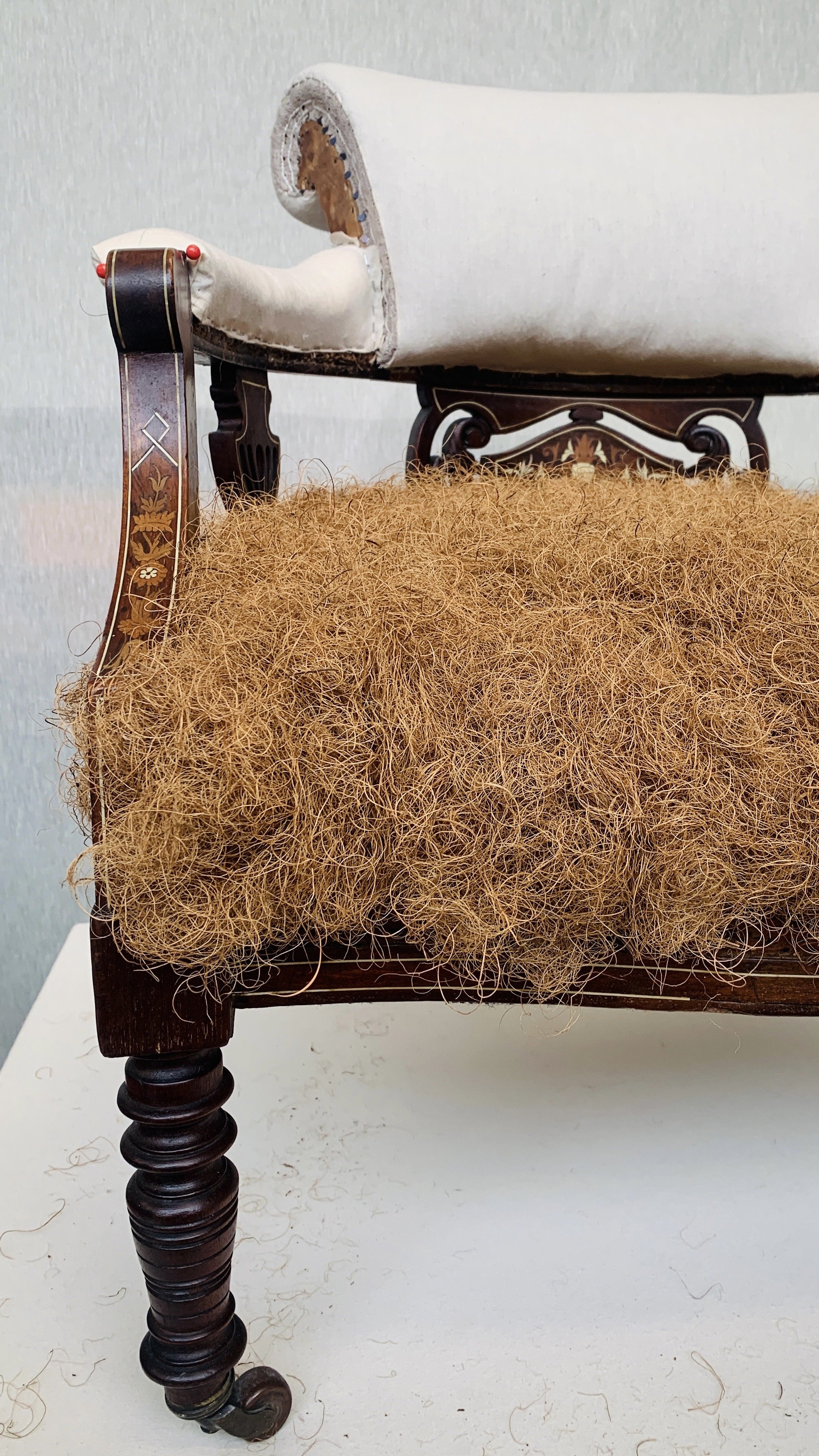 Part of an antique wooden chair with a white upholstered back rest and seat, with a large tangled ball of dried, curly plant fibers resting on the seat.