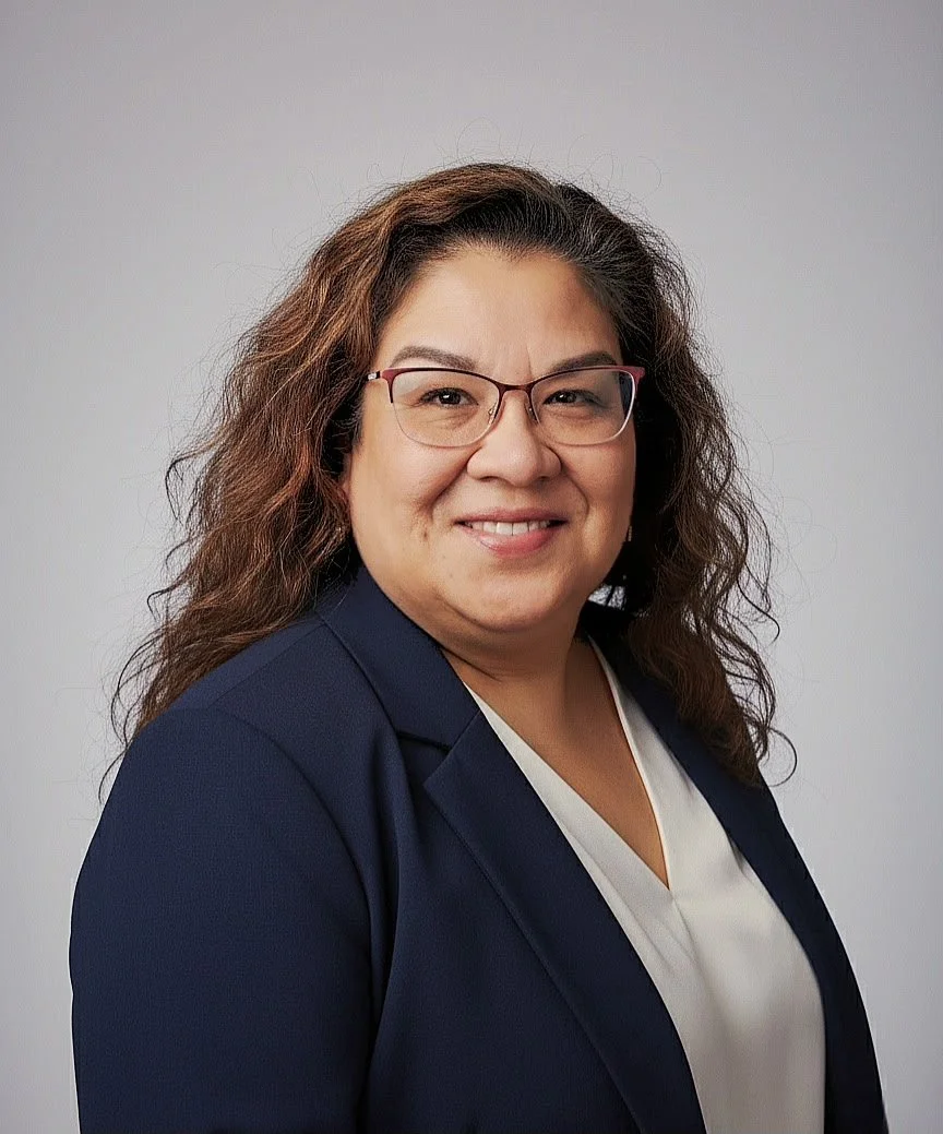 Professional portrait of a woman with glasses, curly brown hair, wearing a navy blazer and a white top, smiling against a neutral gray background.