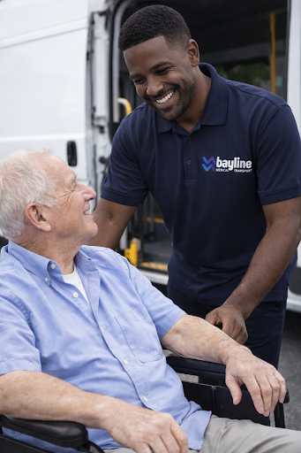 A smiling young man in a navy blue Bayline Transportation polo shirt talking to an elderly man in a wheelchair outdoors.
