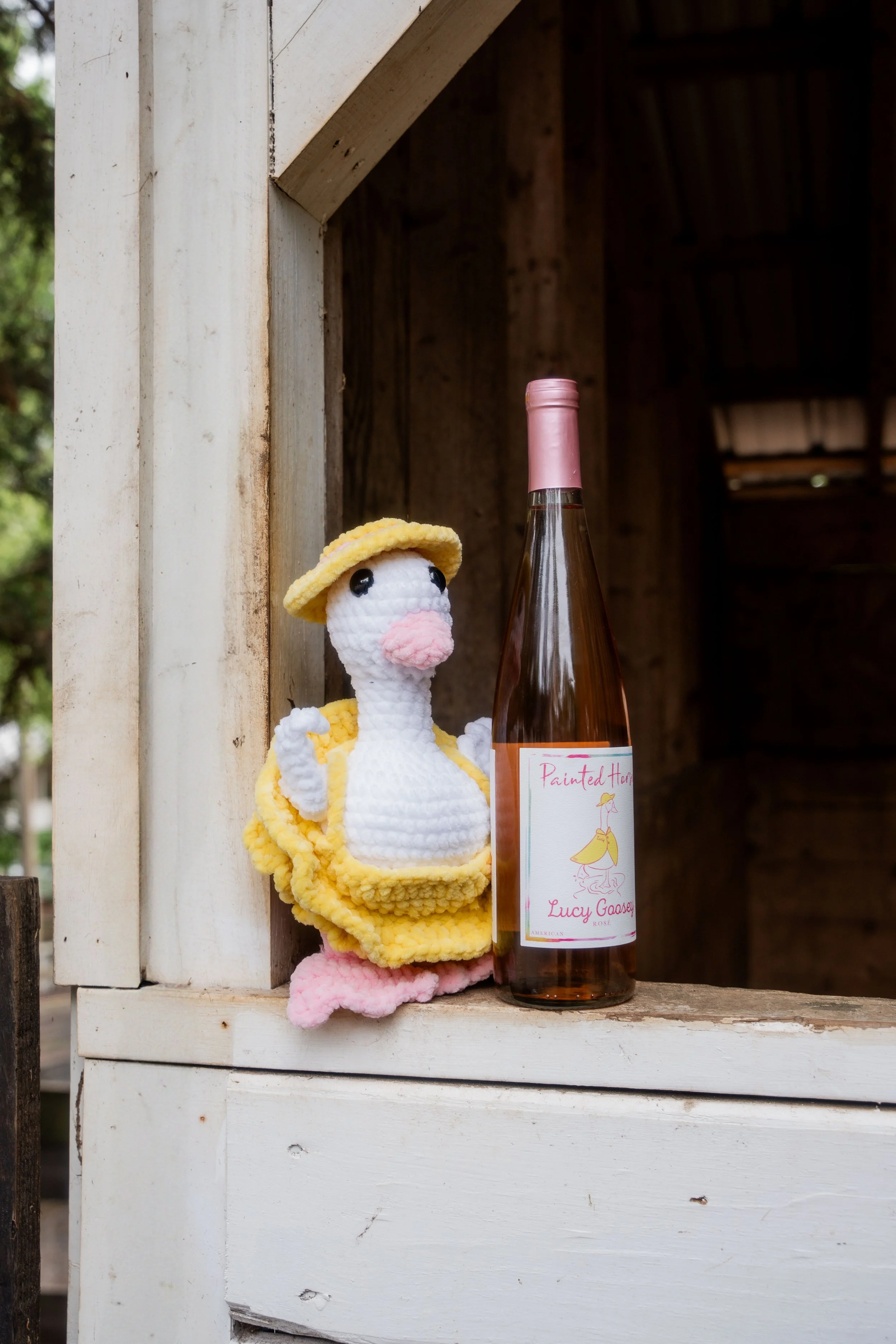 A crocheted duck with a yellow hat and dress, sitting next to a bottle of rosé wine with a handwritten label, placed on a wooden surface outside a rustic wooden structure.
