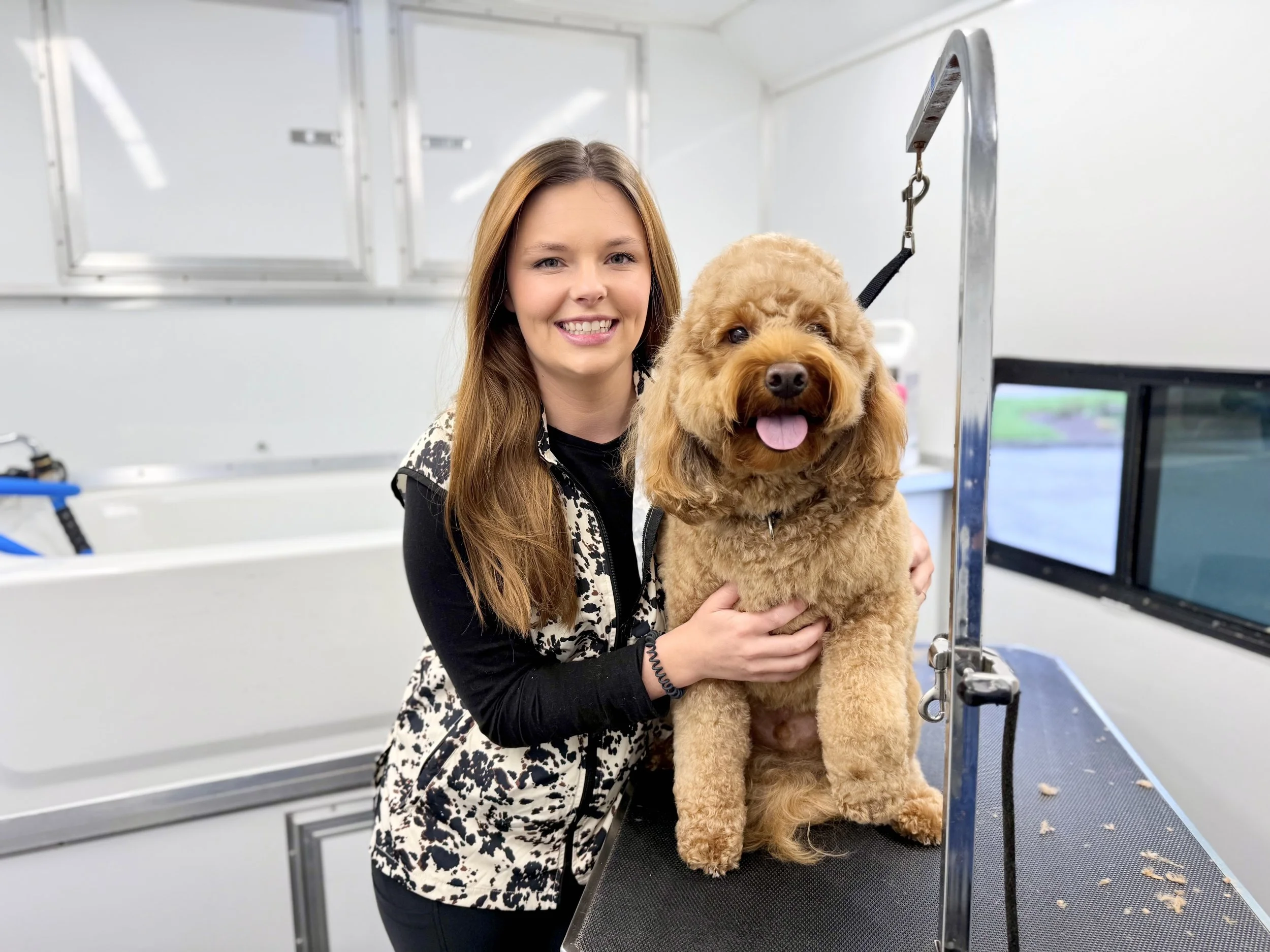 A woman with long brown hair smiling while holding a fluffy brown dog on a grooming table in a pet grooming trailer.