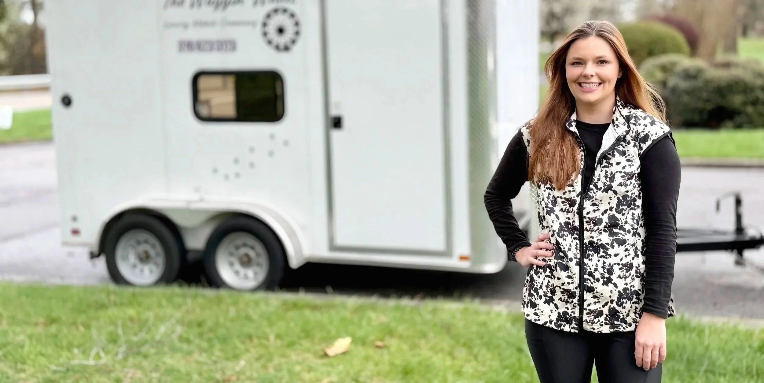 A young woman with long brown hair standing outdoors next to a white trailer, smiling with one hand on her hip.