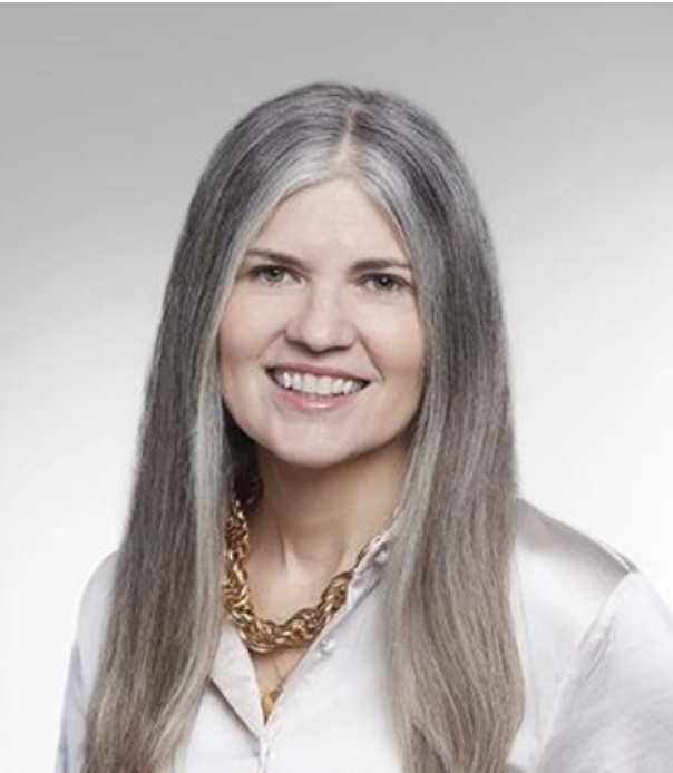 Portrait of a smiling woman with long gray hair, wearing a white shirt and a gold chain necklace, against a neutral background.