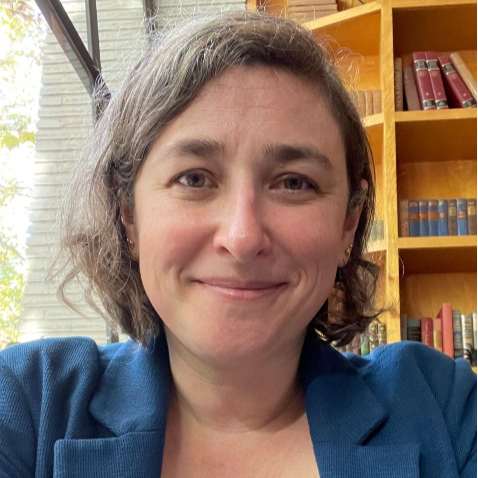 A woman with shoulder-length brown hair smiling in a room with bookshelves in the background.