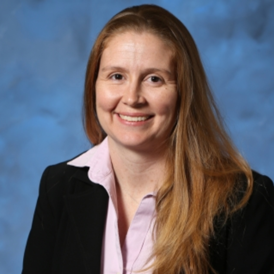 A woman with long red hair smiling, wearing a black blazer and a white collared shirt, against a blue background.