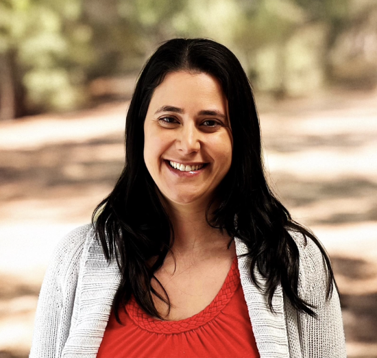 A woman with dark hair smiling outdoors, wearing a red top and a light gray cardigan.