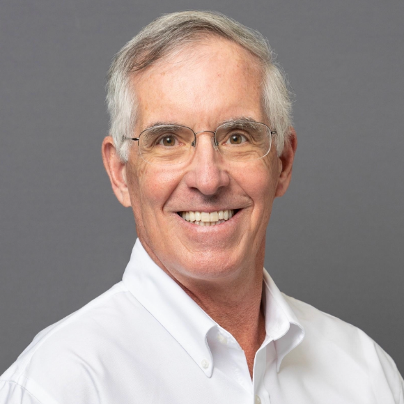 A smiling middle-aged man with gray hair and glasses wearing a white collared shirt against a gray background.
