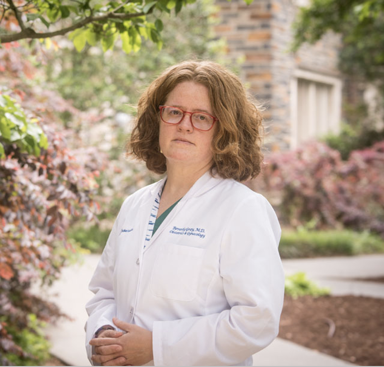 A woman with curly red hair and red glasses standing outdoors in a garden area, wearing a white medical coat.