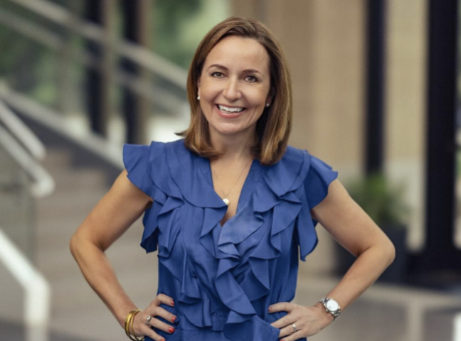 A woman with shoulder-length brown hair smiling, wearing a blue ruffled top and gold jewelry, standing outdoors in front of a building.