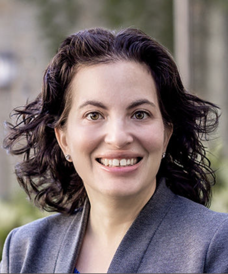 A woman with curly dark hair smiling outdoors, wearing a gray blazer.