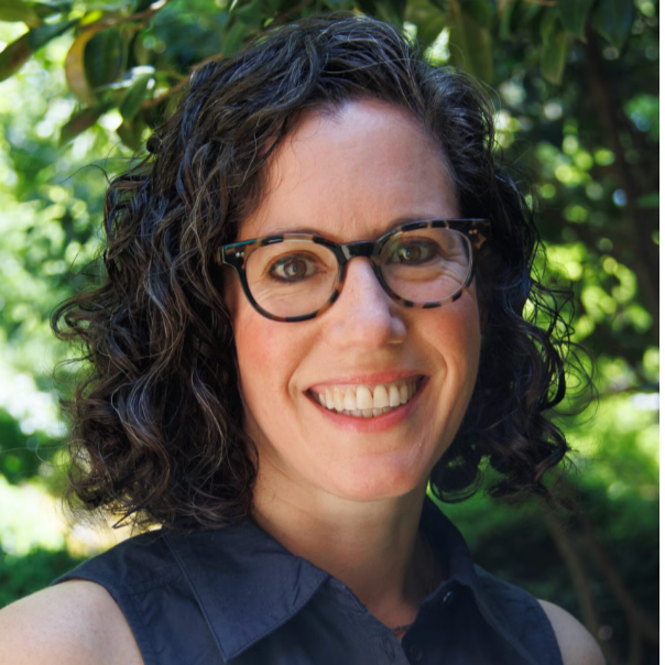 A woman with curly dark hair and tortoiseshell glasses smiling outdoors in front of green foliage.