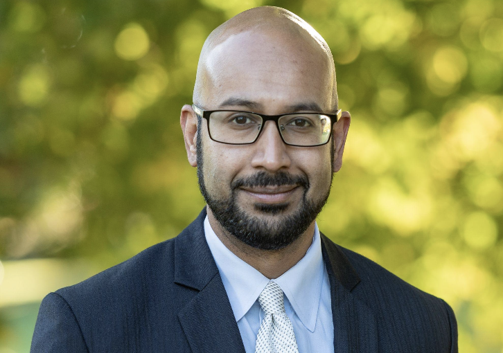 A man with a bald head, glasses, and a beard, wearing a dark suit and light blue shirt with a white tie, standing outdoors with green trees in the background.
