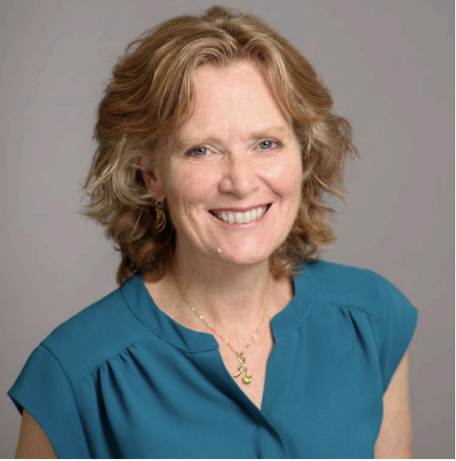 A woman with short curly blonde hair wearing a teal blouse, gold earrings, and a gold necklace, smiling at the camera against a neutral background.