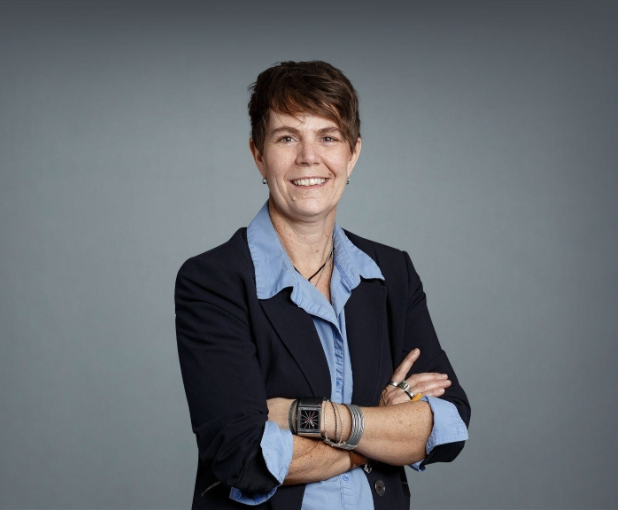 Professional woman with short brown hair, wearing a blue button-up shirt and a dark blazer, smiling with arms crossed against a gray background.