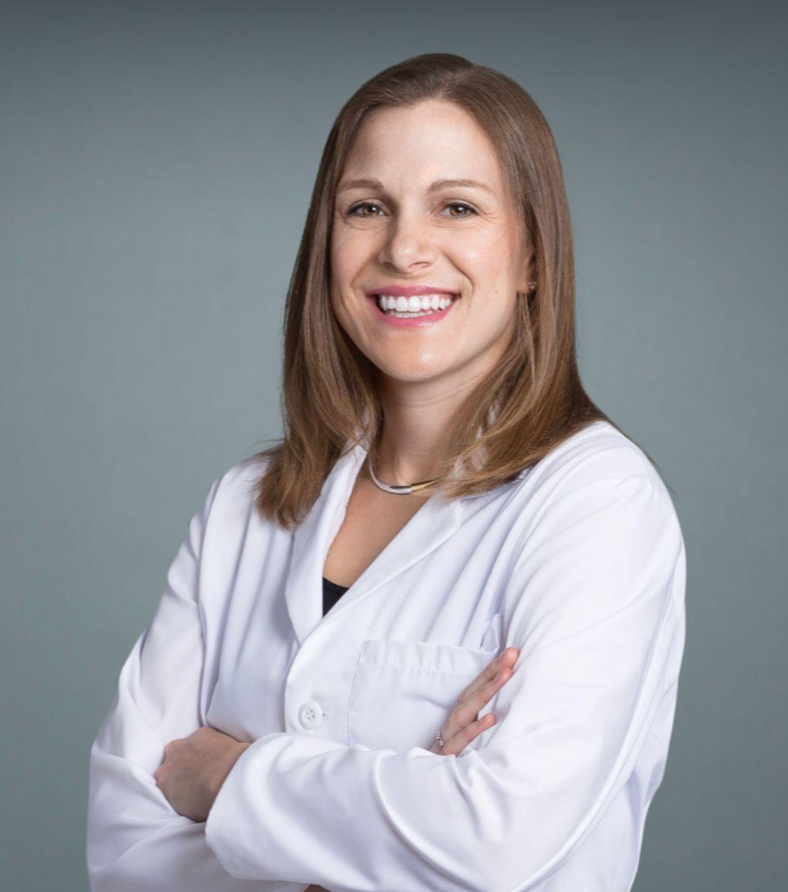 A woman with light brown hair, smiling, wearing a white medical coat, standing with her arms crossed against a neutral background.