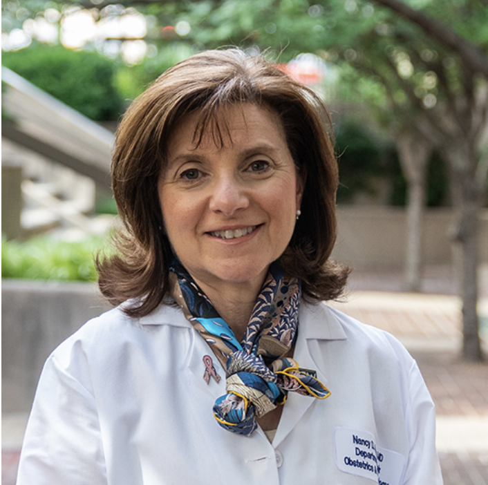 A woman in a white medical coat with a patterned scarf tied around her neck, smiling outdoors with greenery and trees in the background.