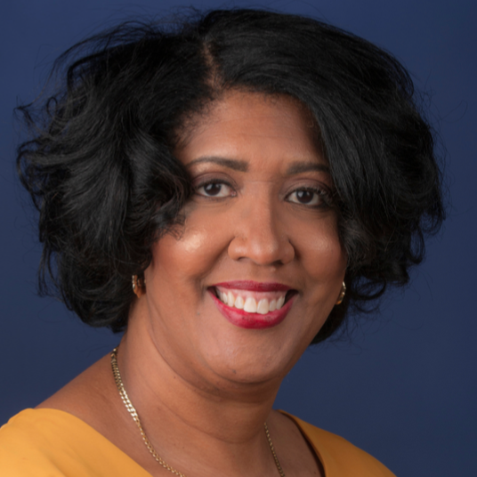 A woman with black, curly hair dressed in a yellow top smiling against a blue background.