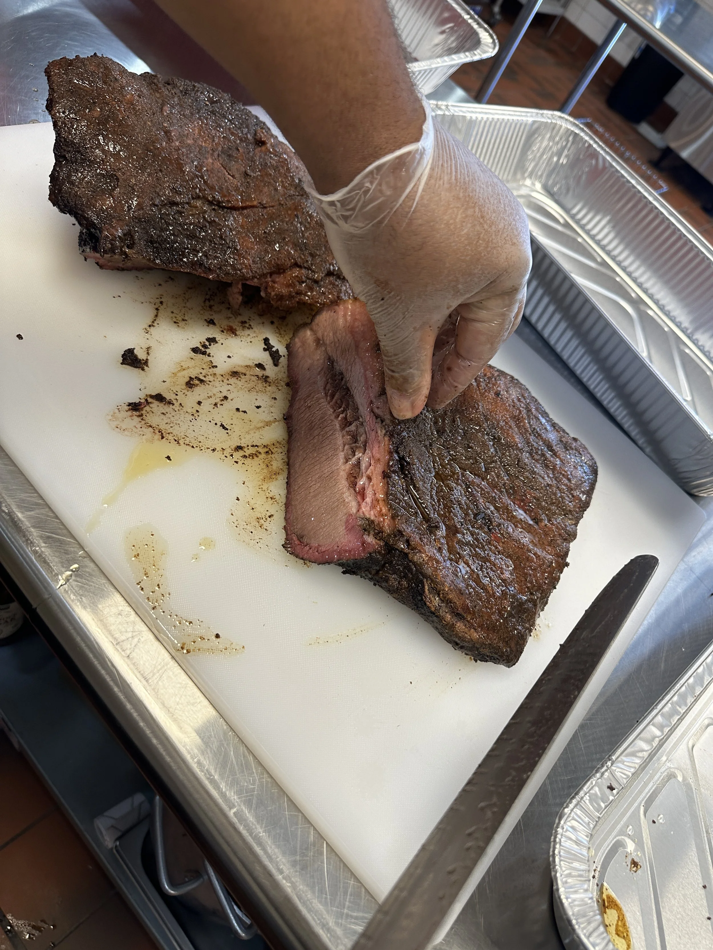 A person cutting a smoked beef brisket on a white cutting board, with disposable gloves, several aluminum trays, and a knife nearby, in a kitchen or catering setting.