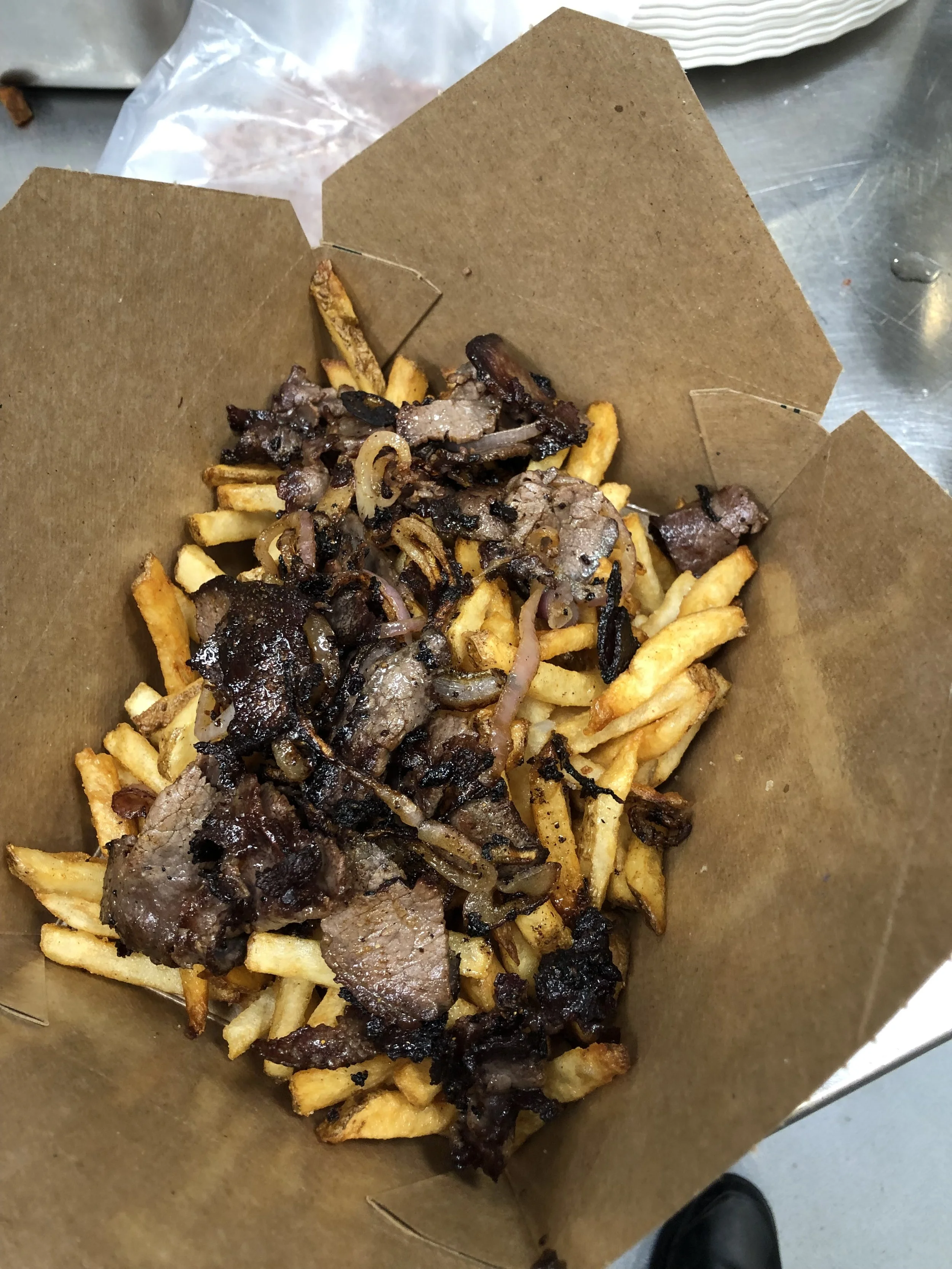 A serving of French fries topped with cooked chopped beef, grilled onions, and dark gravy in a brown paper food basket.
