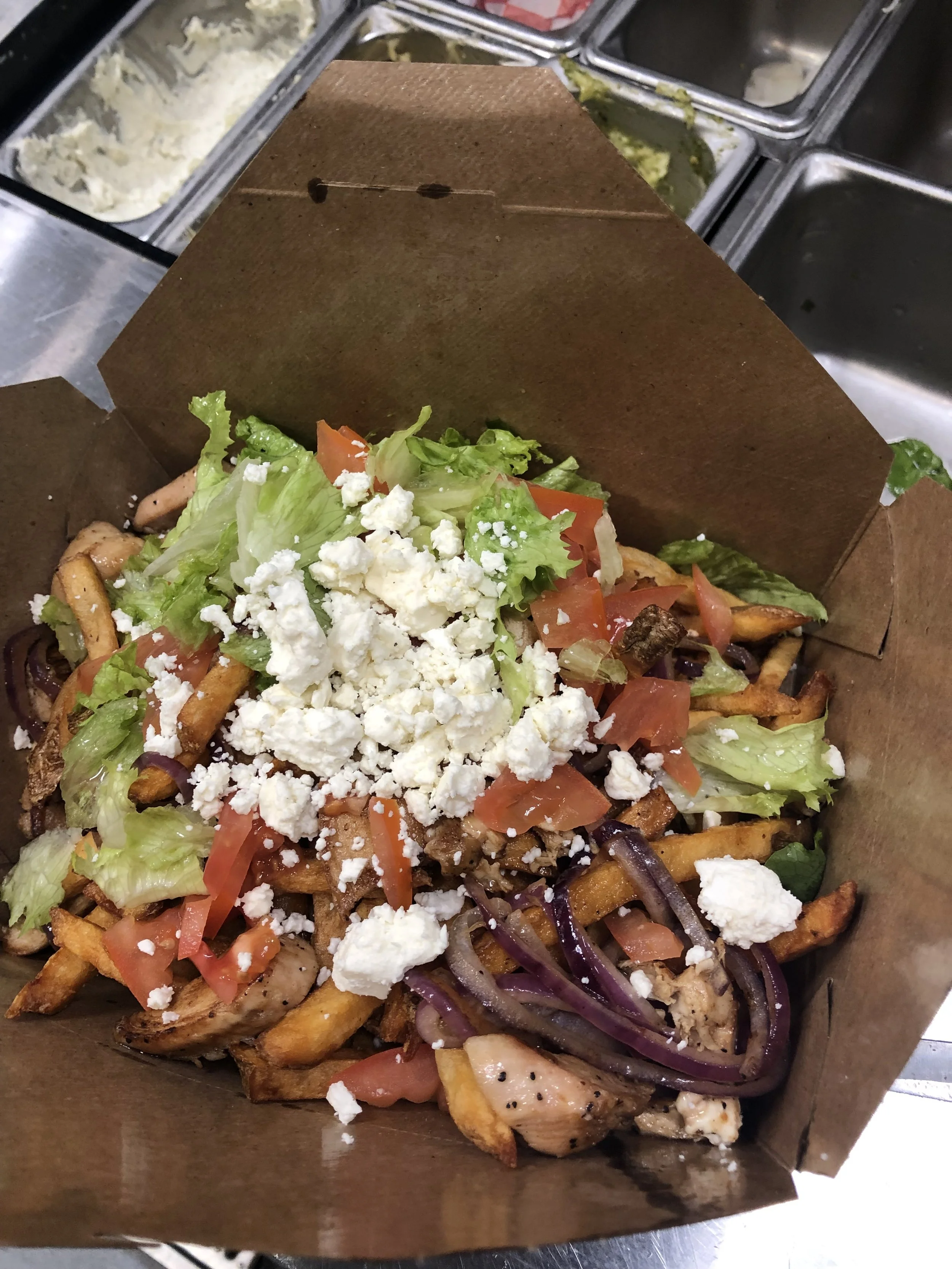 French fries topped with chopped tomatoes, onions, grilled chicken, shredded lettuce, feta cheese, and other vegetables inside a paper food tray.