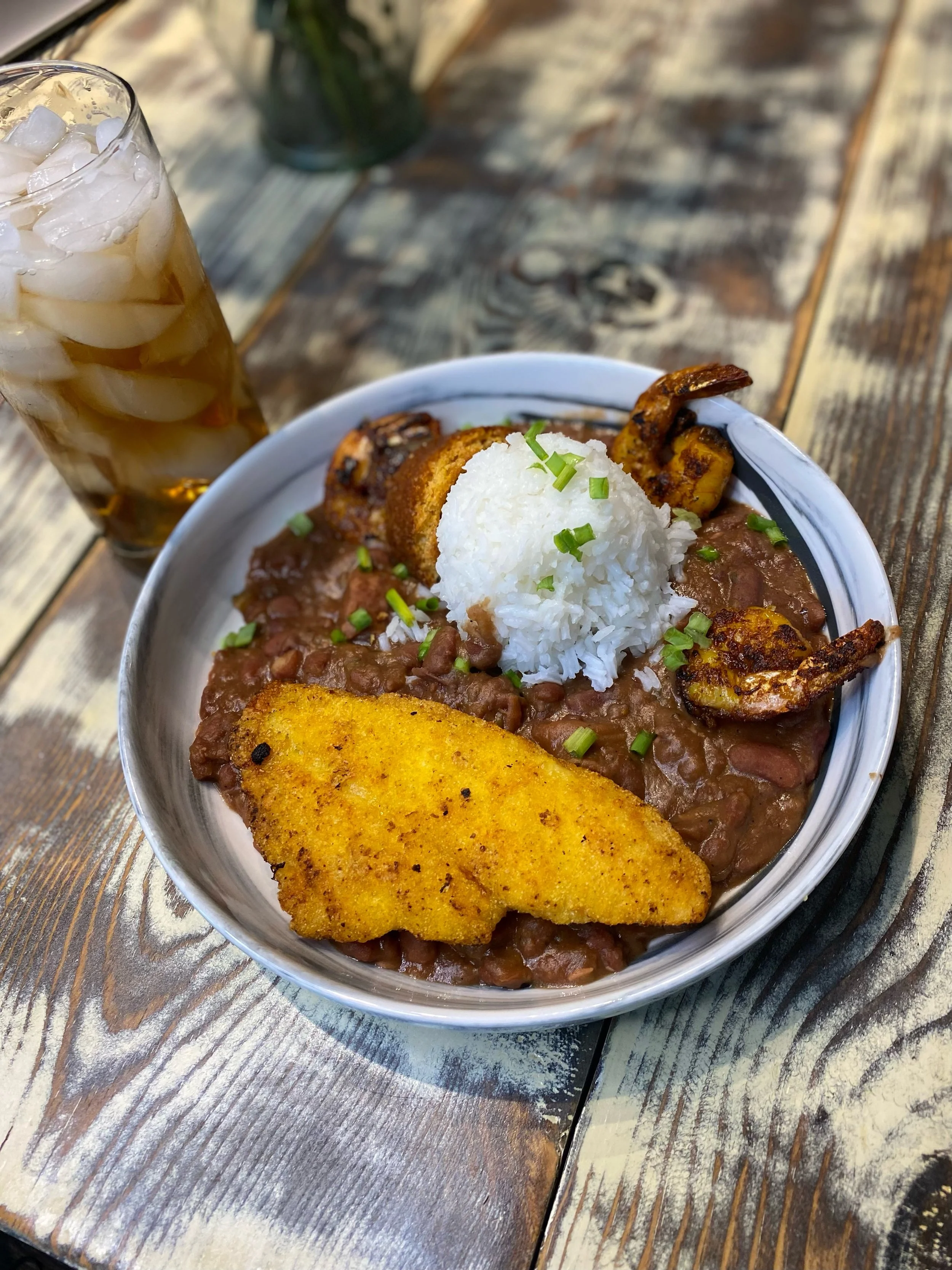 A bowl of rice, fried plantains, running beans, and grilled shrimp meal on a wooden table with a glass of iced tea nearby.