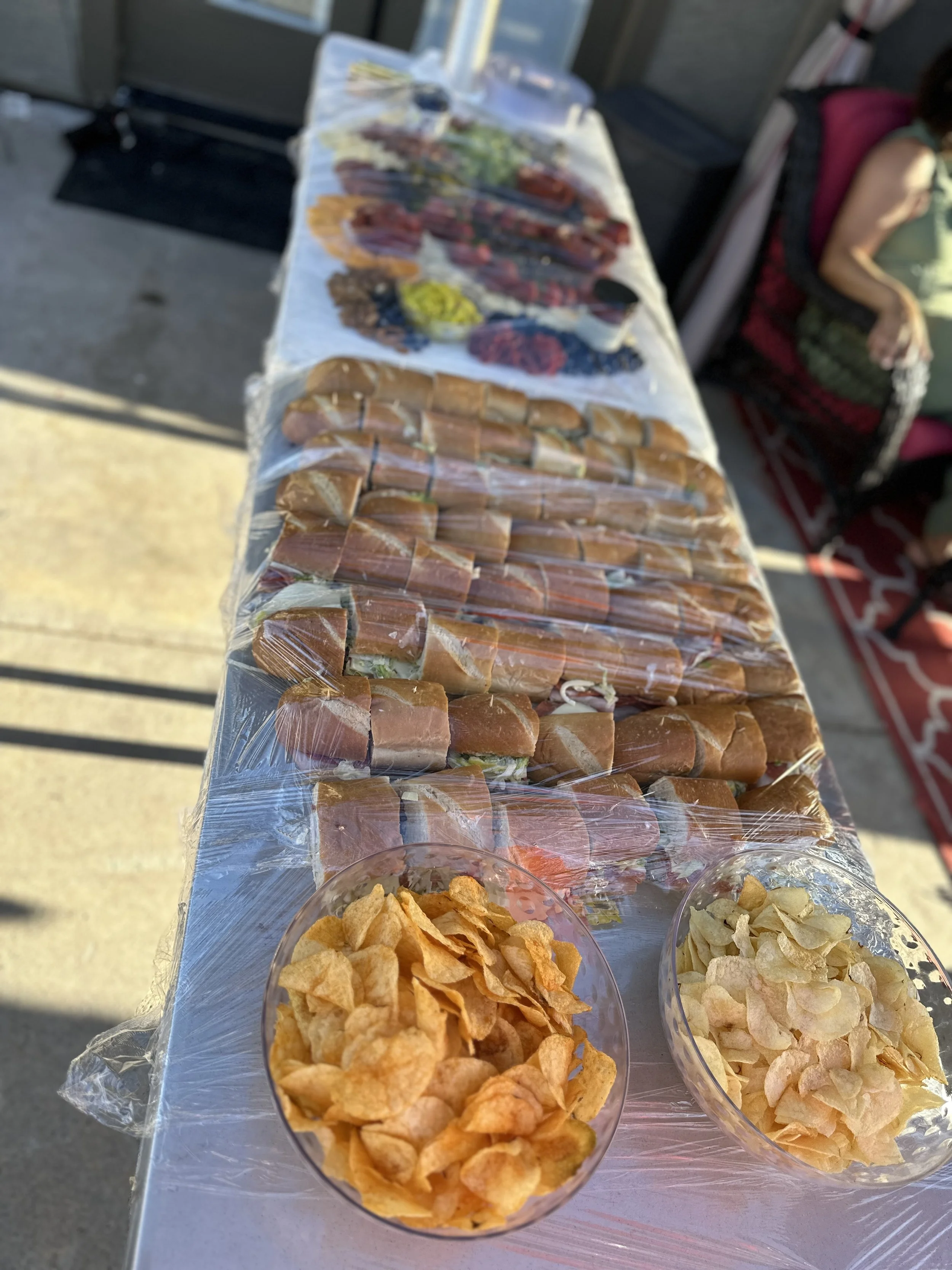A display of food on a table including wrapped sandwiches, bowls of potato chips, and a platter of sliced meats and vegetables, at an outdoor event.