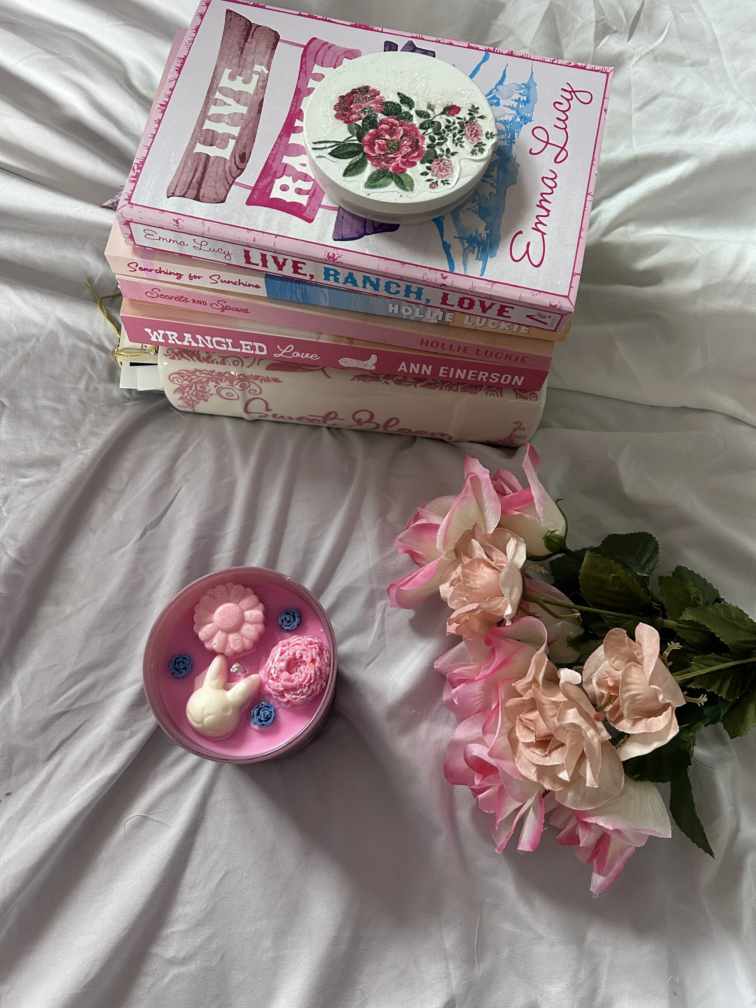 Stack of five pink and white books with floral design on top, a pink and white candle with lei and bunny-shaped chocolate inside, and pink roses on a white bedsheet.